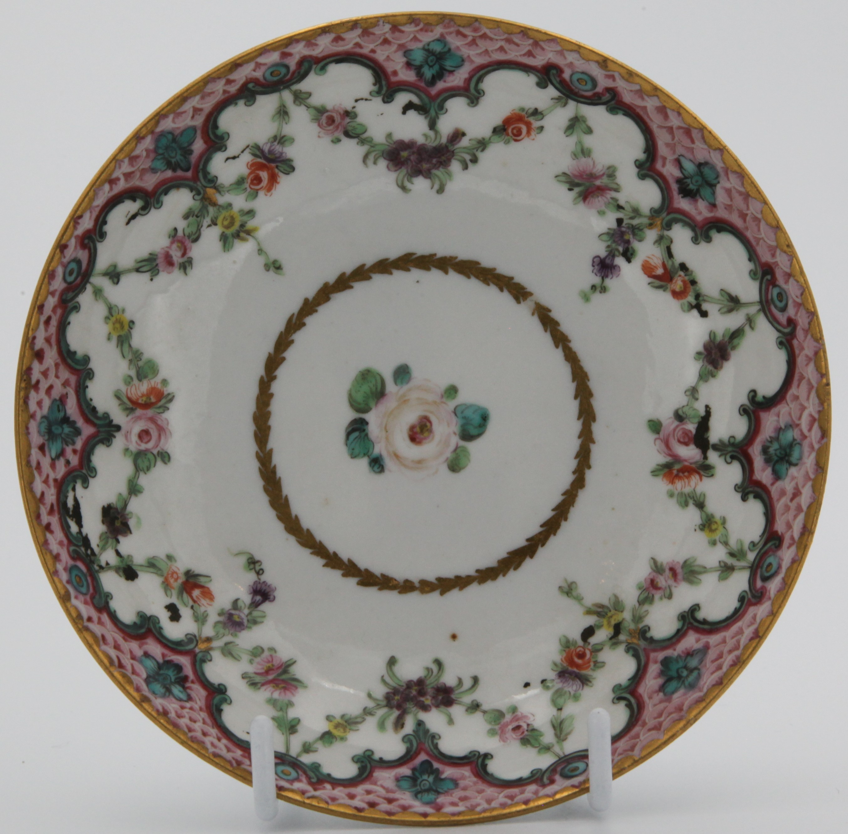 A tea saucer from a rare Bristol cabaret set, the cup with double ‘C’ scroll handle, painted in coloured enamels with scattered flowers, within a pink scale border, with green ‘C’ scrolls and flower heads, suspending garlands of flowers and leaves,  circa 1775.