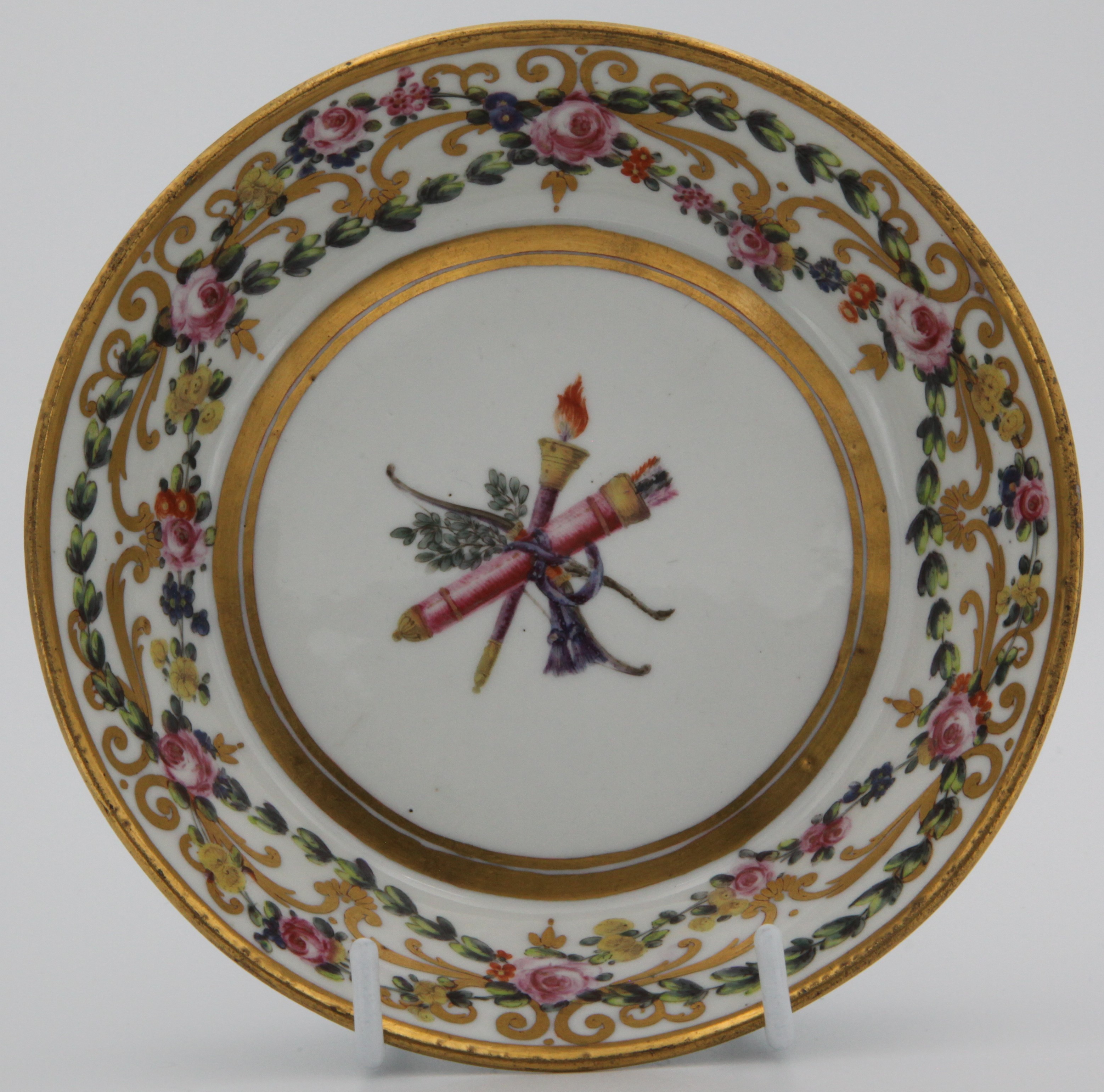 An extremely fine Bristol cabinet saucer, superbly painted in coloured enamels with swags of flowers, and roundels of pink roses suspended from blue ribbons, within gilt leaf scrolls, the saucer with a central trophy emblematic of Love, circa 1775, blue painted X marks and numeral 3. in gilt