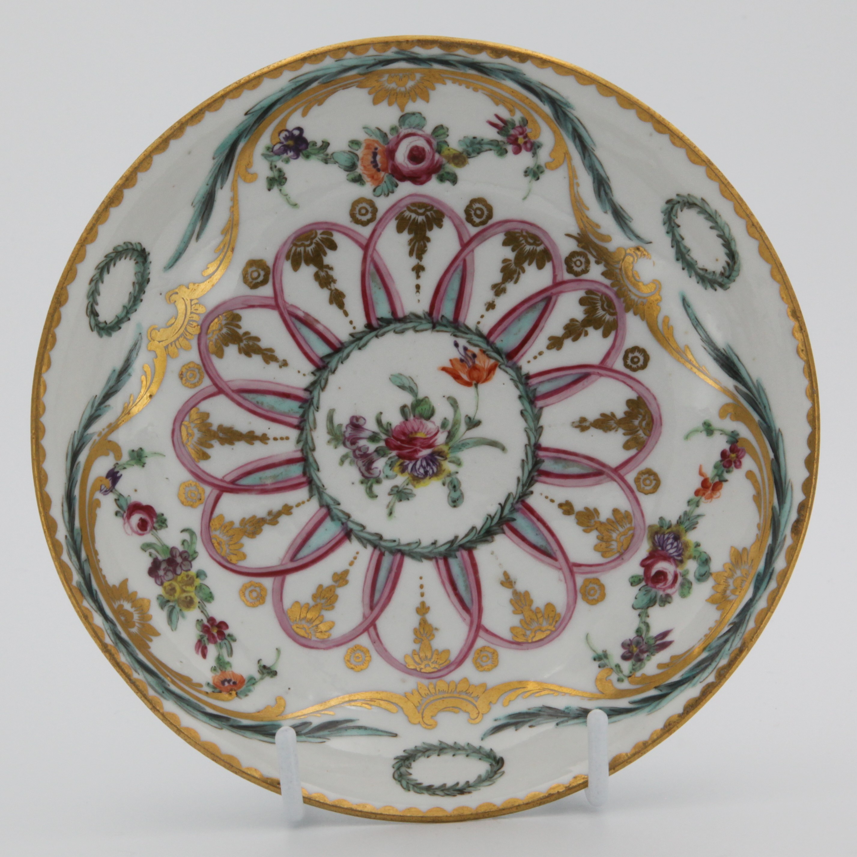 A very fine Bristol tea bowl saucer, painted in coloured enamels with flowers and leaves, within scrolling gilt leaf and green and turquoise laurel bands, with oval laurel garlands, and with a pale blue, puce and gilt interlined band, gilt dentil rims, circa 1775, blue painted crossed swords marks and numeral 2. in gilt