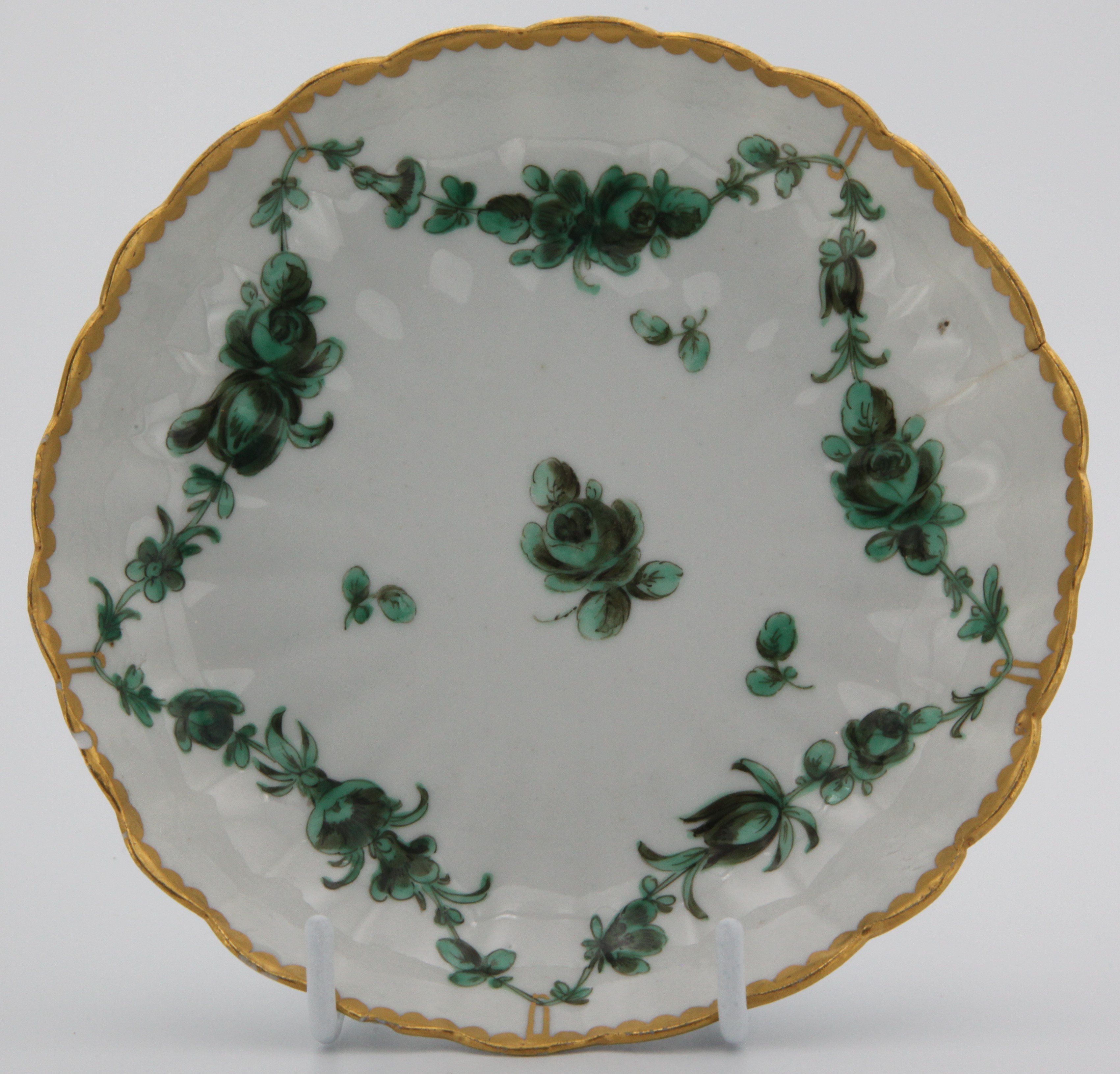 A fluted Bristol porcelain saucer, the wavy rim which is richly gilded. The decoration is in green and black. It comprises a single central flower surrounded by a continuous garland suspended from the rim at five equidistant points by gilded hoops. Mark: blue X 1.