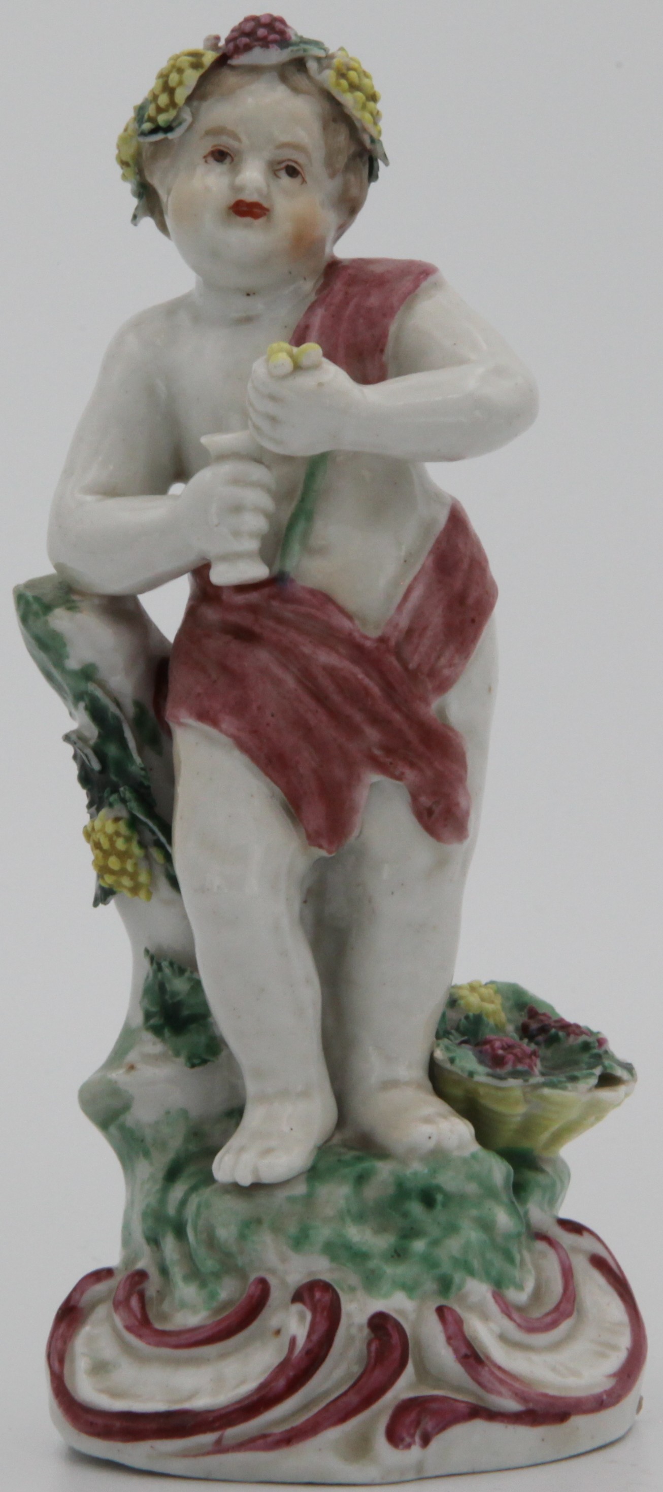 A Plymouth figure of Bacchus representing Autumn from the Infant Four Seasons. The figure is of a putto holding some grapes and a wine glass while leaning on a stump with a basket full of grapes by his side. Rococo scroll work to the moulded mound base highlighted in puce. The figure is in superb condition with some chipping to the leaves as shown. No mark