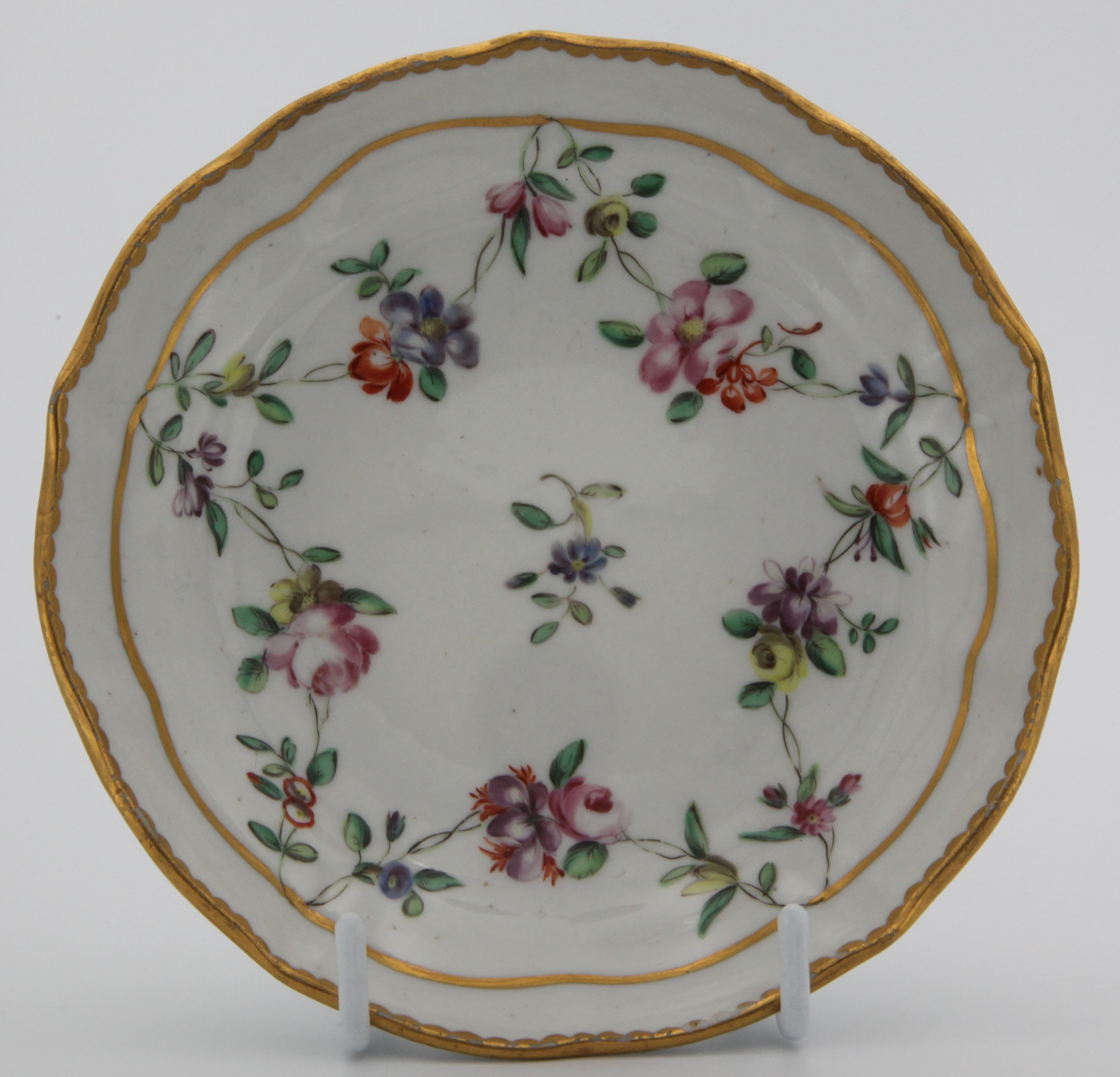 A Bristol saucer, slightly lobed, painted in enamel with garlands of colourful flowers suspended from a heavy gilt line around a central blue flower spray, surrounded by heavy gilt dentil rim. Mark: blue X5.