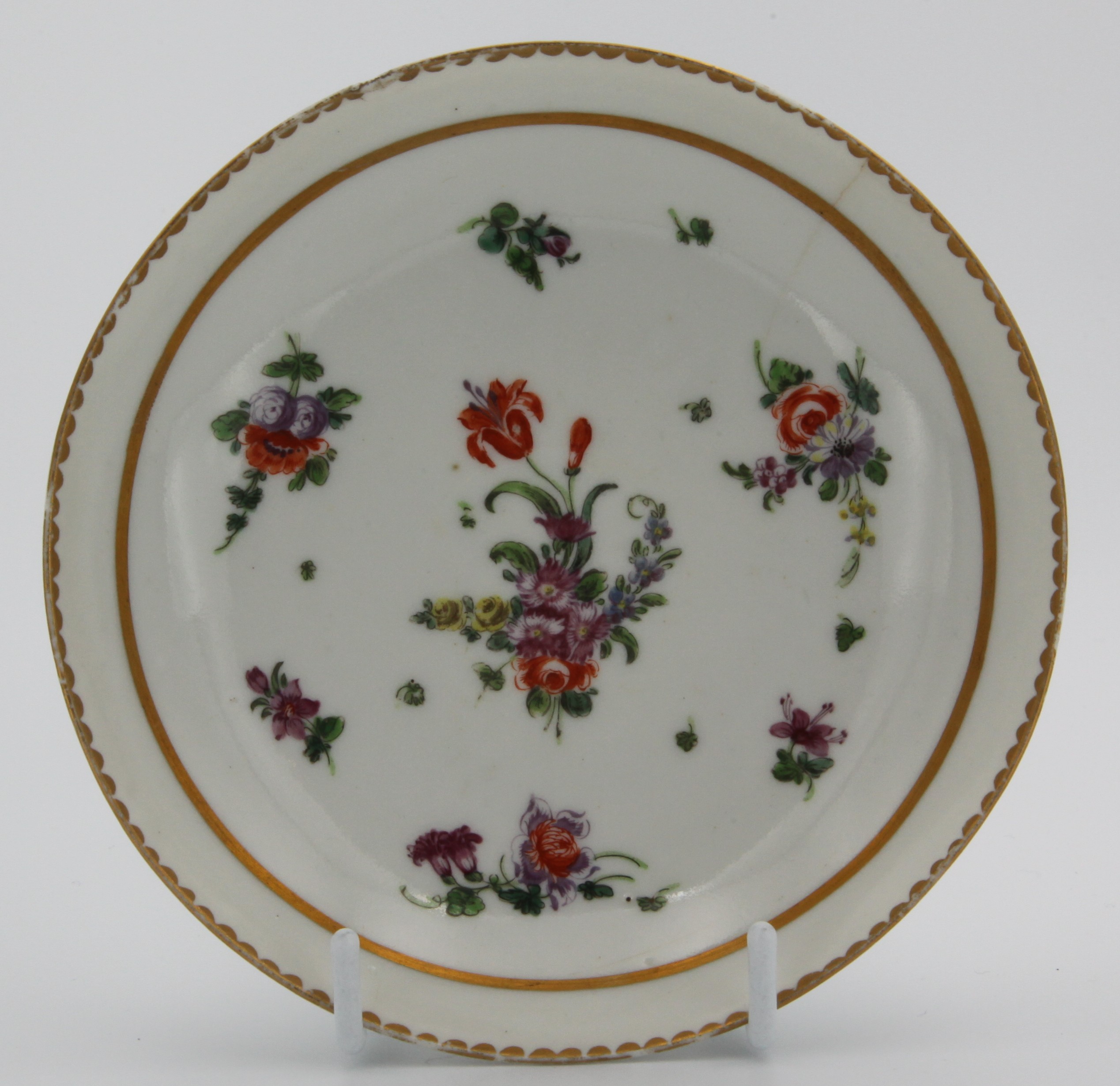 A Bristol tea saucer, painted in colourful enamels with a central spray with prominent orang flower and scattered sprays and leaves inside a thick gilt band, surrounded by gilt dentil rim, c.1770. No mark