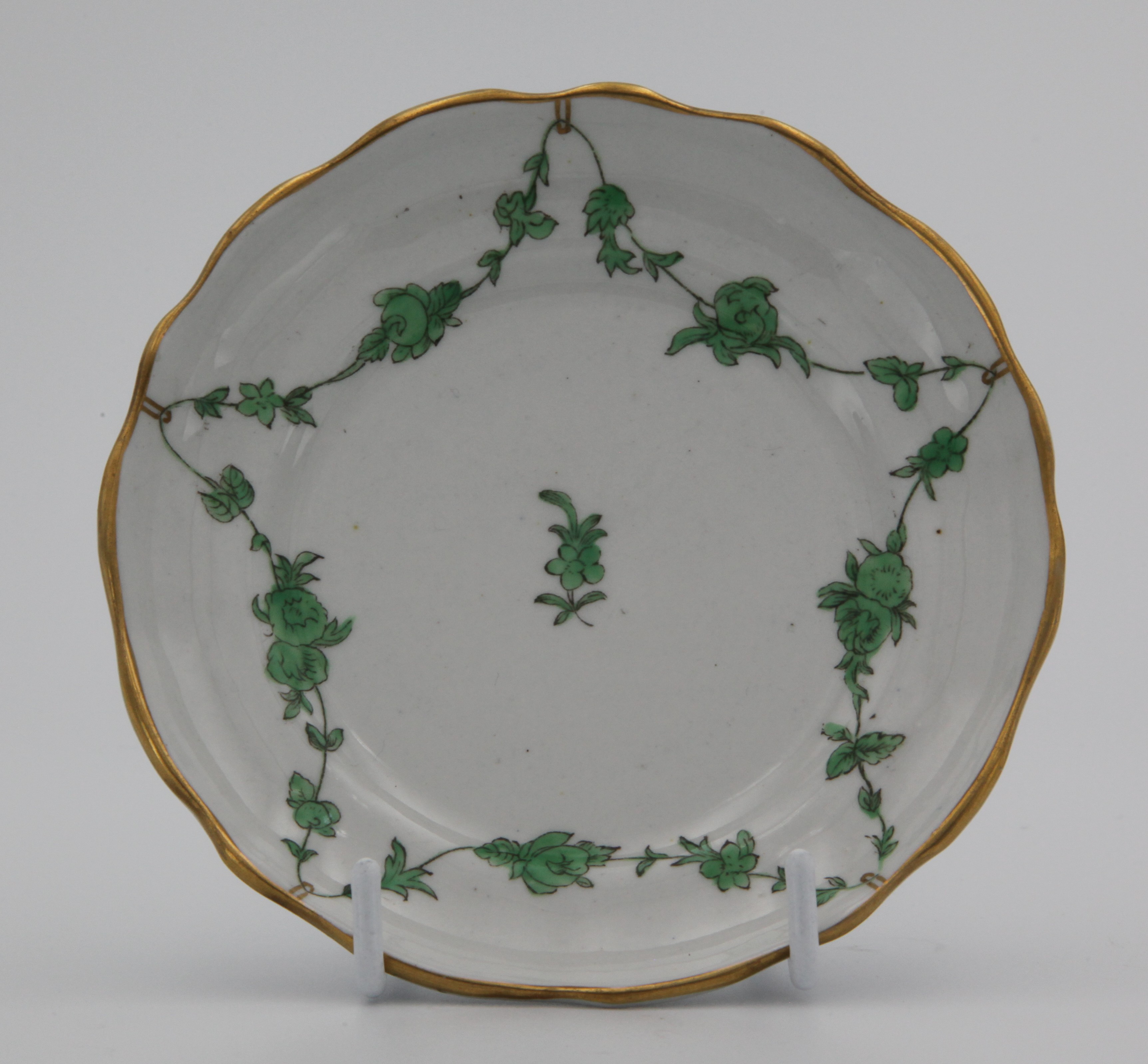 A Bristol lobed, ogee-shaped tea saucer painted with green monochrome garlands of flowers and leaves suspended from gilt loops on gilt rim. Blue X10 mark