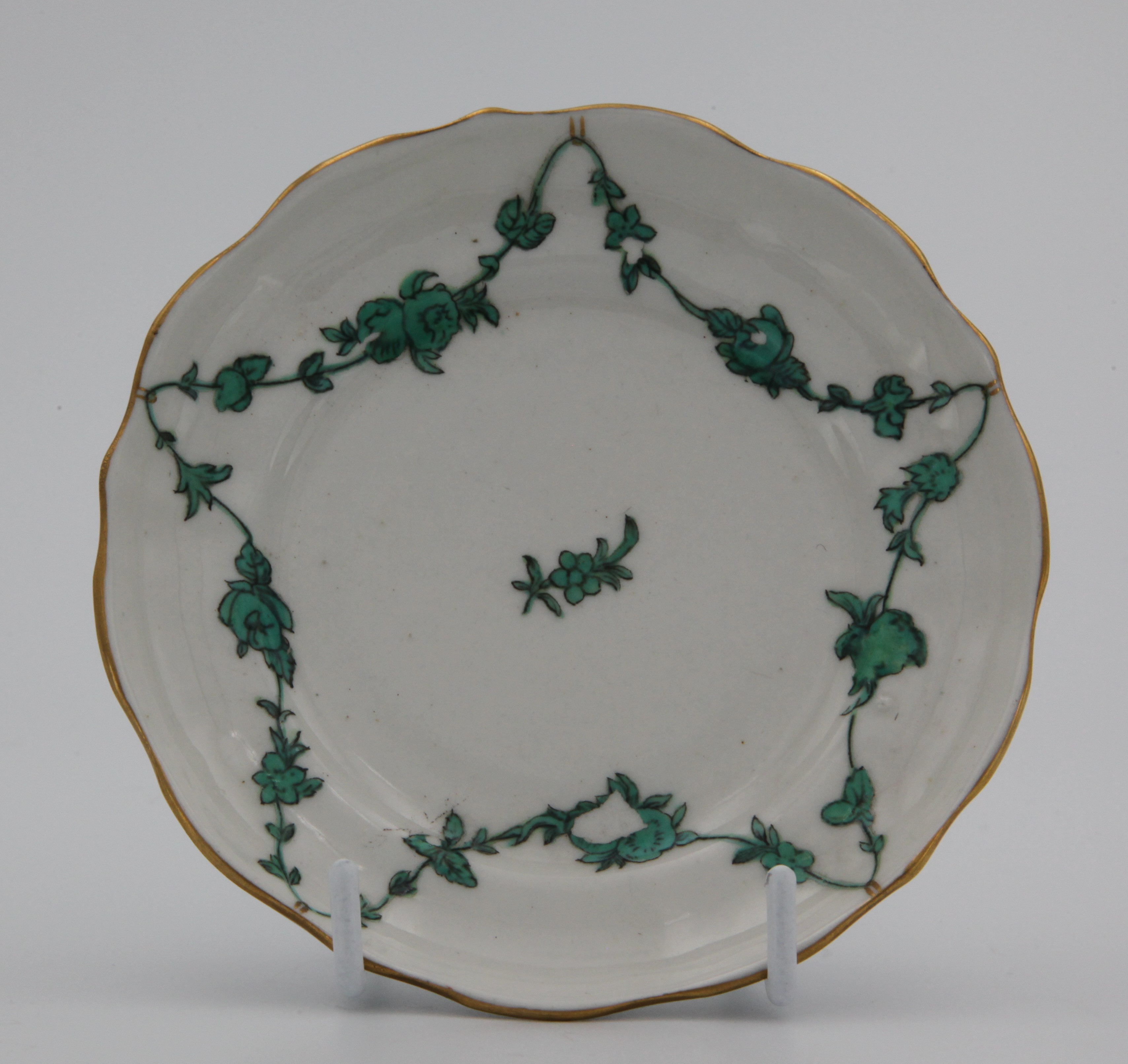 A Bristol lobed, ogee-shaped tea saucer painted with green monochrome garlands of flowers and leaves suspended from gilt loops on gilt rim. Blue X10 mark