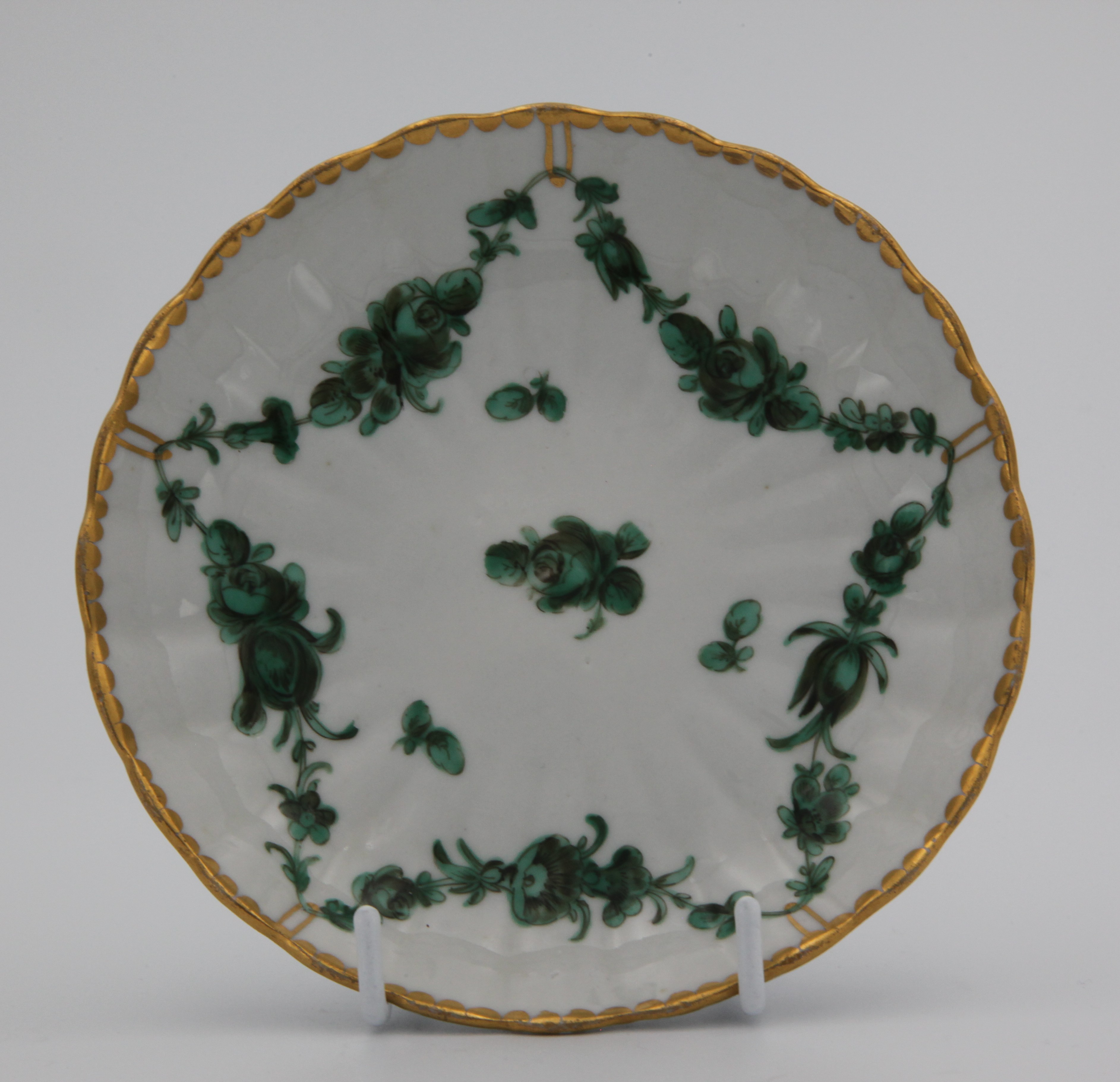 A fluted Bristol porcelain saucer, the wavy rim which is richly gilded. The decoration is in green and black. It comprises a single central flower surrounded by a continuous garland suspended from the rim at five equidistant points by gilded hoops. Mark: blue X1.