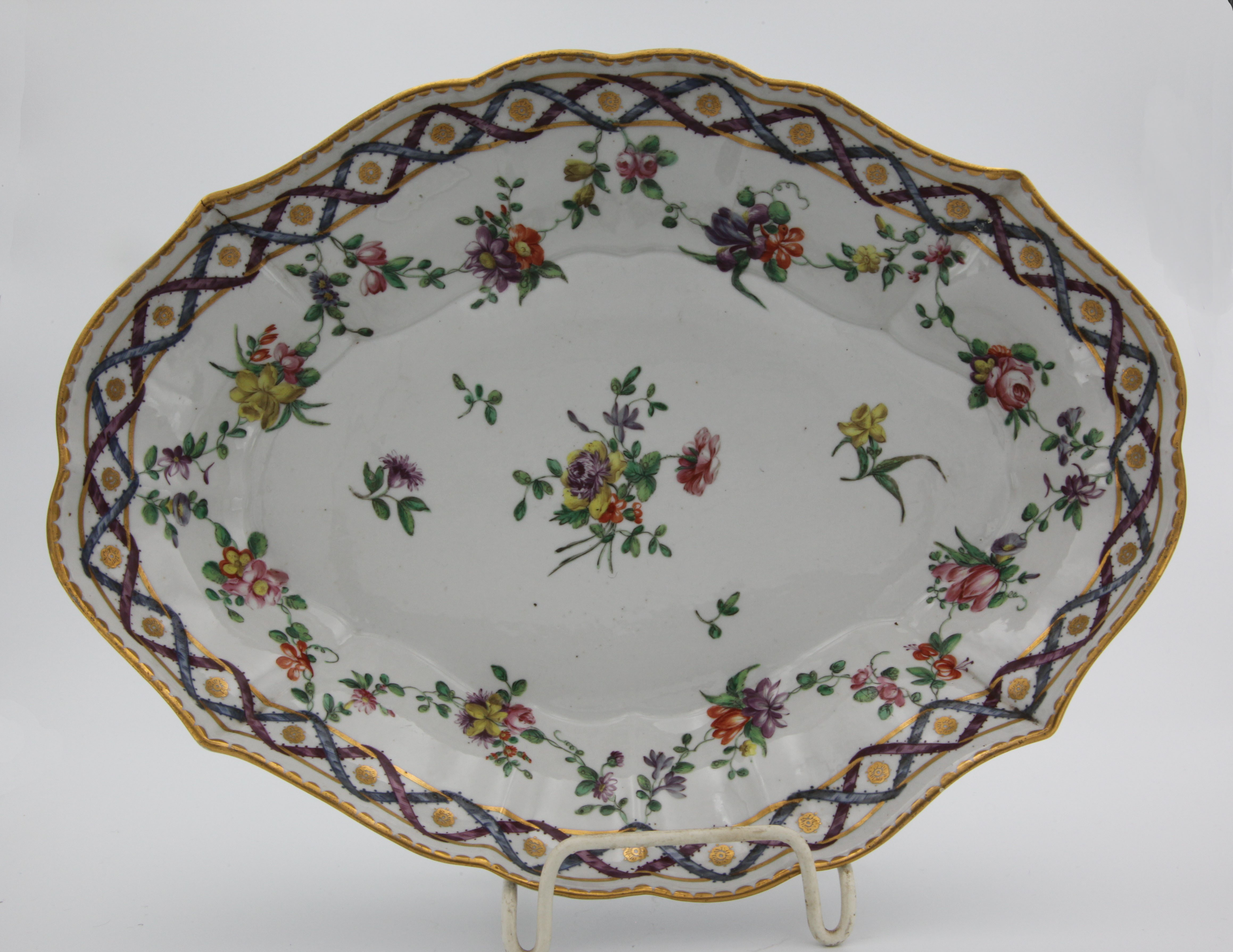 A Bristol fluted lozenge-shaped dish, painted in colourful enamels with a flower spray and scattered flowers, the puce and blue interlocked border with gilt flower head medallions, and suspending swags of flowers and leaves, gilt dentil rim, 10" wide, circa 1775, blue painted X mark. Condition: perfect. See Albert Amor, The Alfred Trapnell Collection, 1912, numbers 170-181, for a part dessert service in this pattern