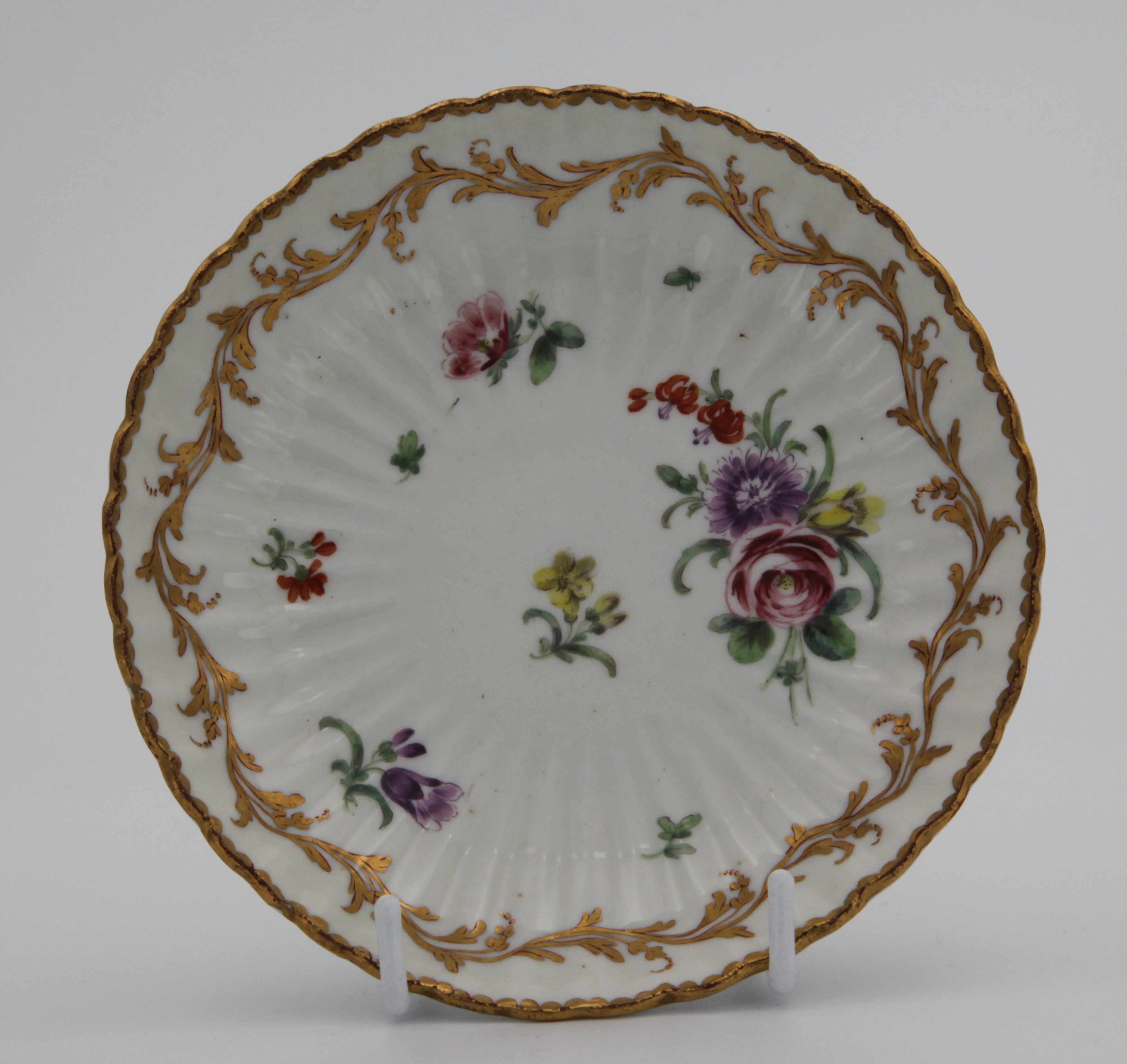 A fine fluted Bristol coffee saucer, painted in enamels with a spray of flowers and scattered leaves and individual flowers, and with an elaborate leafy gilt band, mark blue X1. gilt.