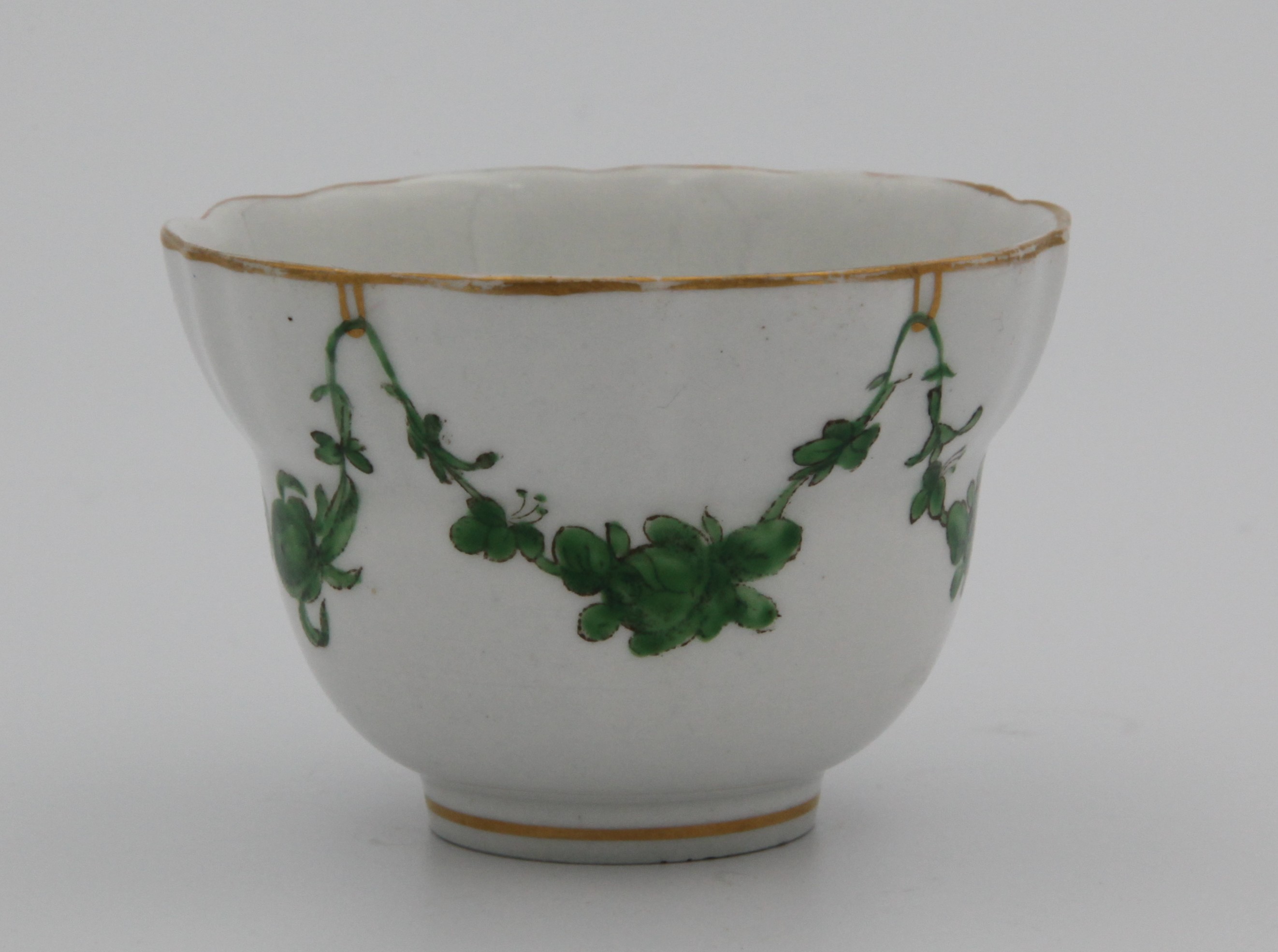 An ogee-shaped Bristol tea bowl, slightly fluted, painted in enamels with monochrom green garlands suspended from gilt looops hanging from a gilt rim line. Mark blue X10