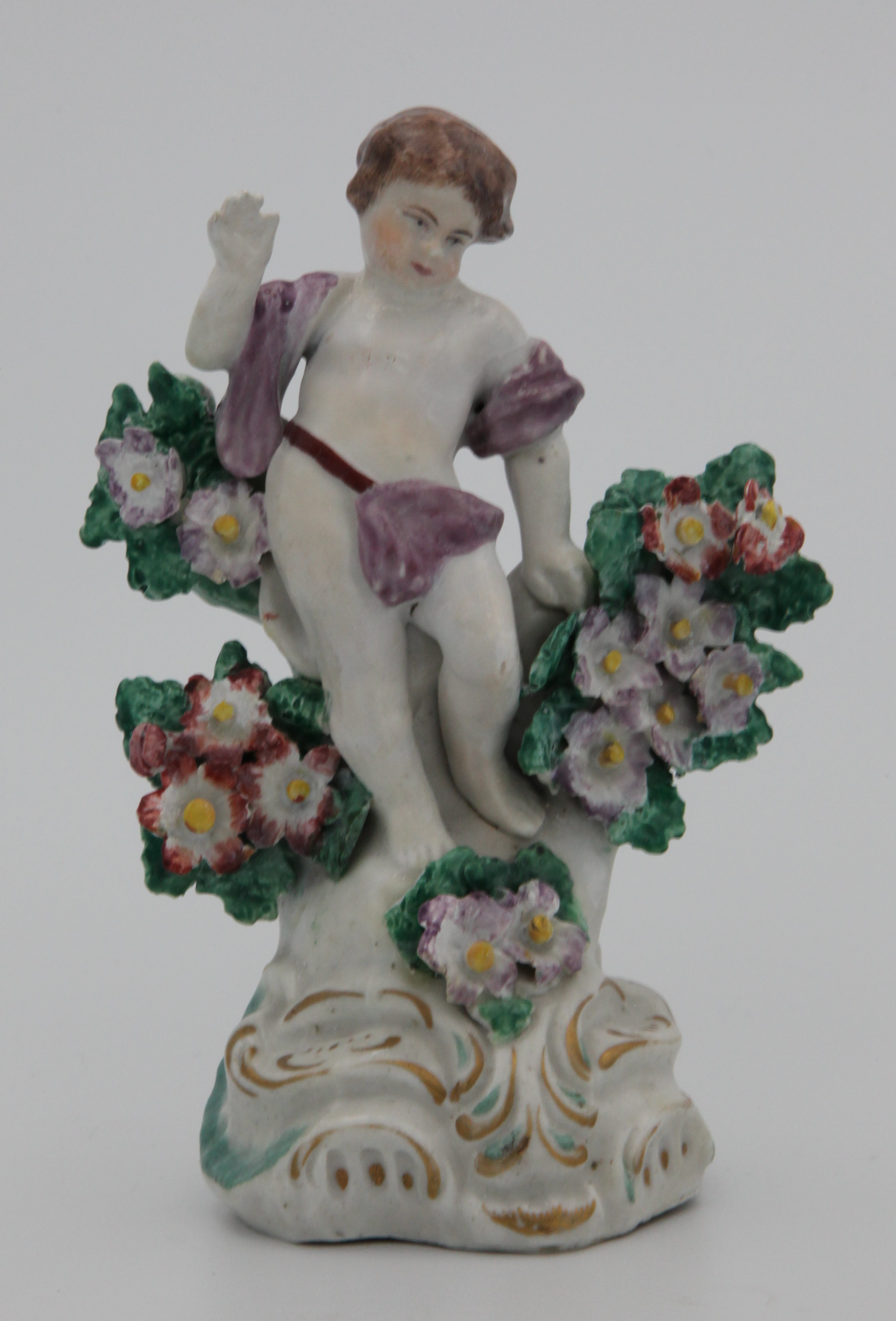 A Plymouth figure of a putto, standing, his right arm raised, and wearing a puce sash, and flanked by floral bocage, the scroll moulded mound base picked out in turquoise and gilt, circa 1768-70, no mark