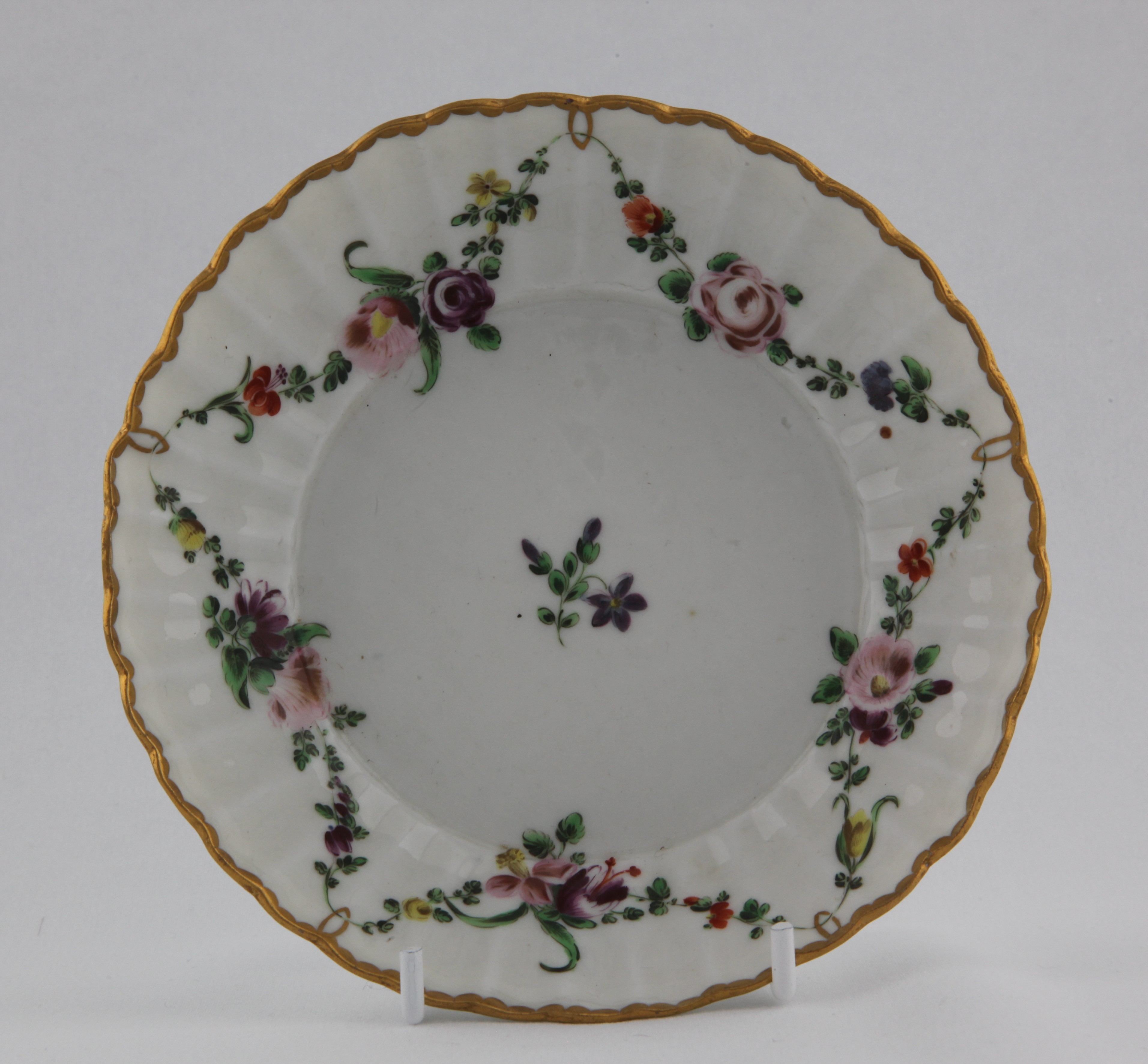 Saucer, from the Gilt loop garland service, decorated by No mark, A fluted Bristol saucer, painted in enamels with a central spray and garlands of colourful flowers and leaves suspended from gilt loops hanging from a gilt dentil rim — Plymouth & Bristol porcelain piece 1482