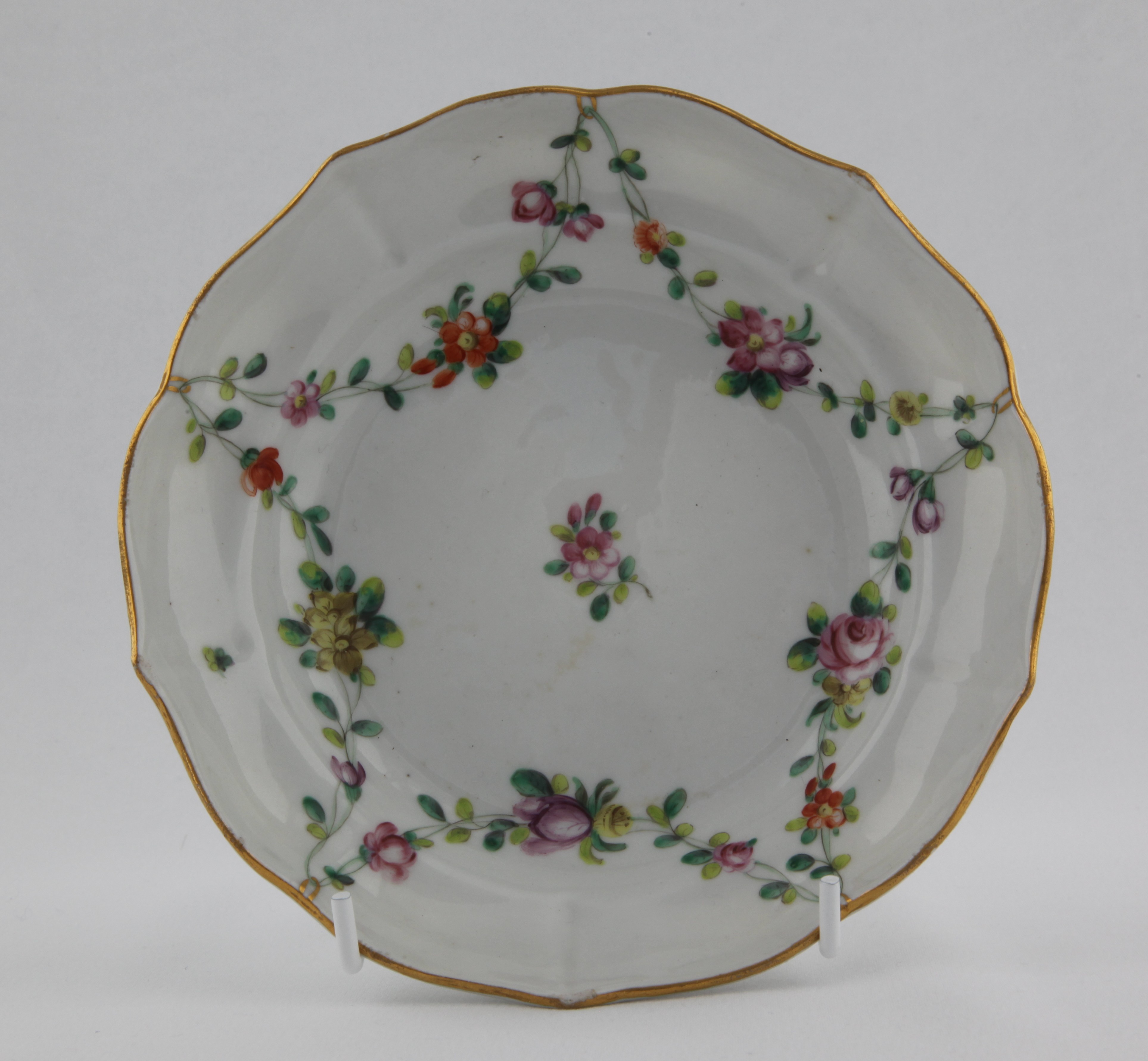 Saucer, from the Gilt loop garland service, decorated by No mark, A Bristol saucer, ogee-shaped and mildly lobed, painted in enamels with garlands of colourful flowers and leaves suspended from gilt loops hanging from a gilt rim line — Plymouth & Bristol porcelain piece 1484