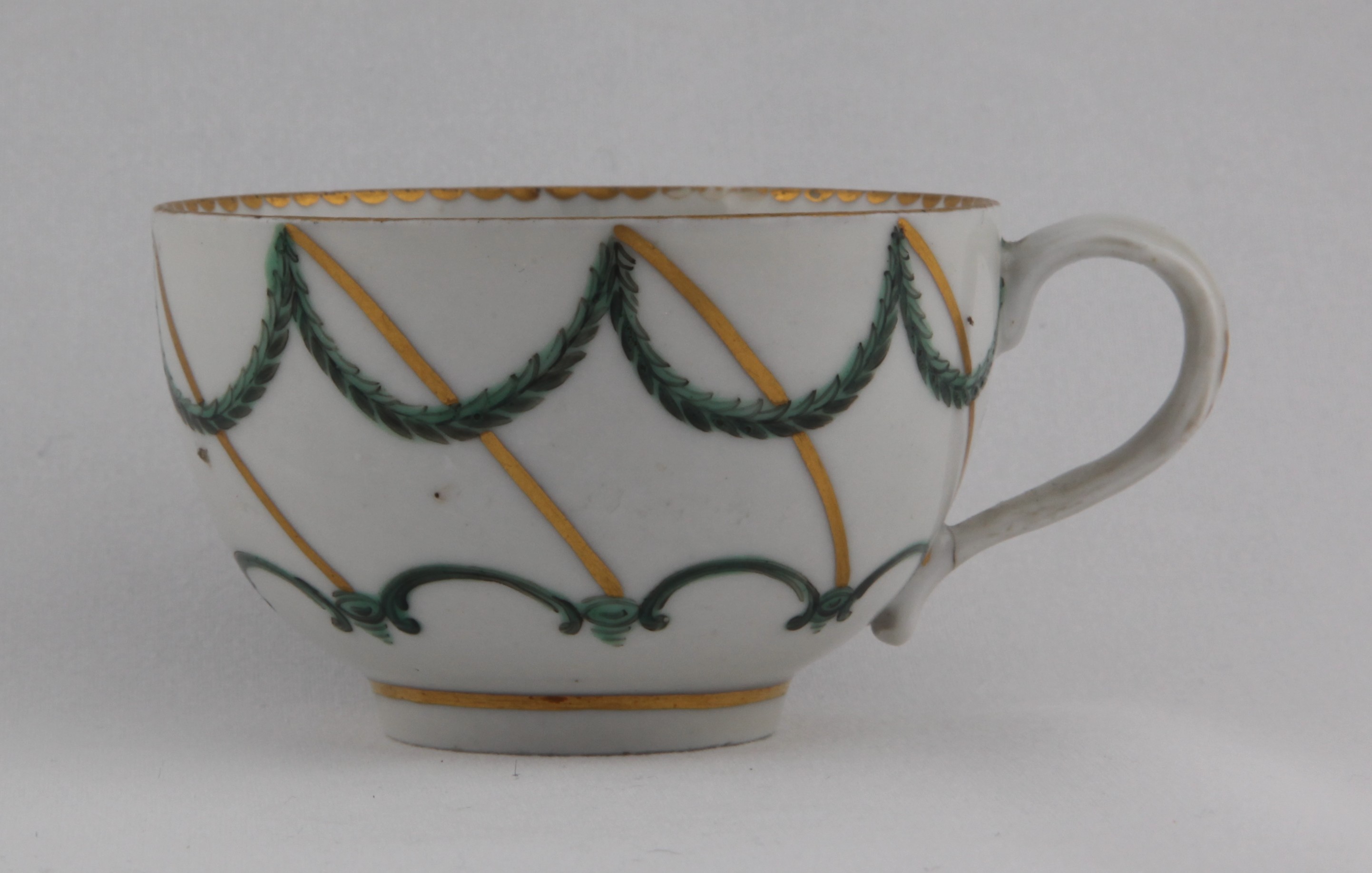 A fine Bristol tea cup of rounded shape with moulded loop handle, painted in enamels with a lower green looping band below green laurel swags suspended from diagonal gilt lines, gilt dentil rim to the inside