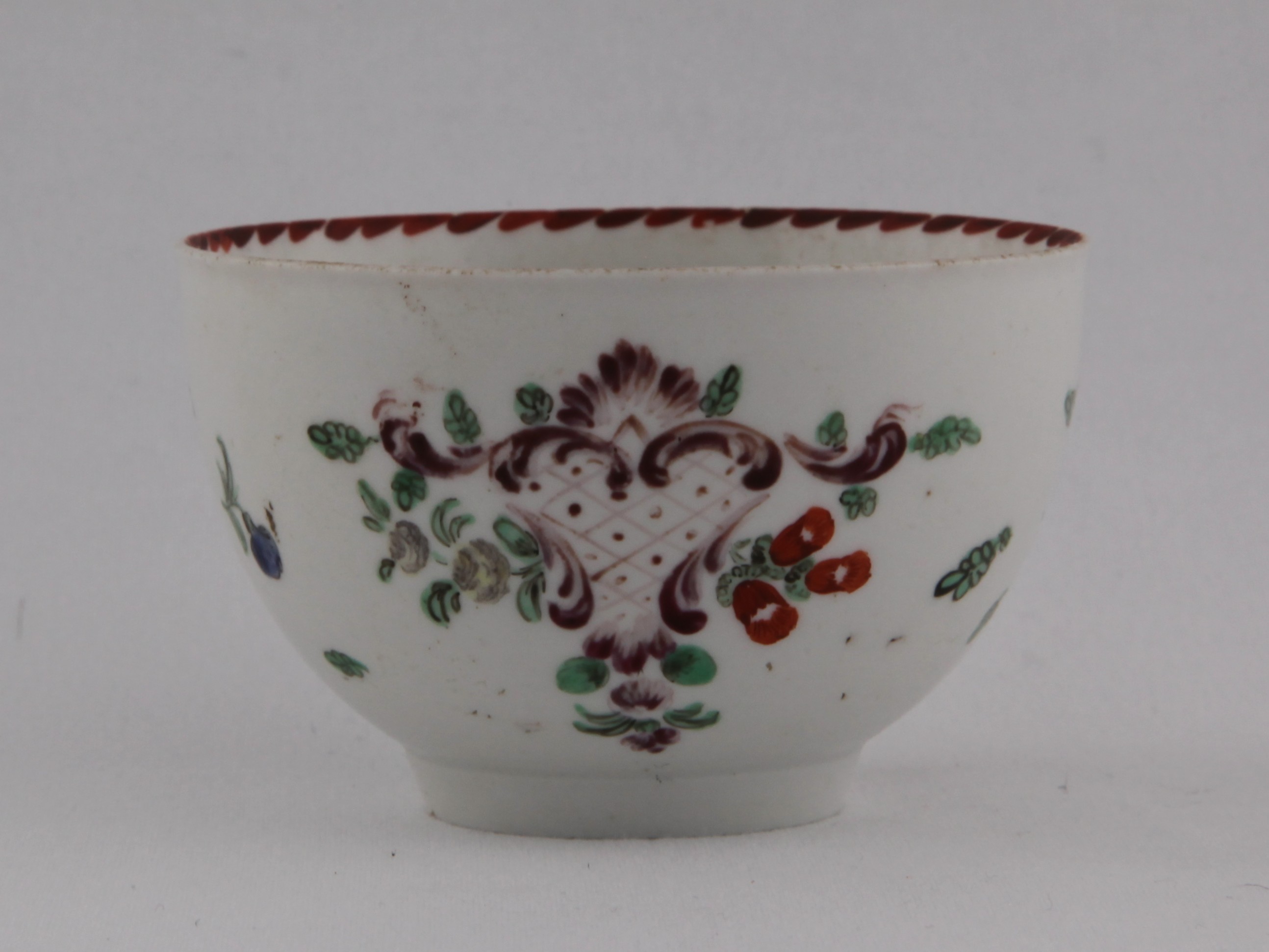 A Bristol tea bowl of slightly everted rounded shape painted in enamels with a puce cartouche surrounded by scattered flowers and leaves, and orange angular dentil rim line to the inside