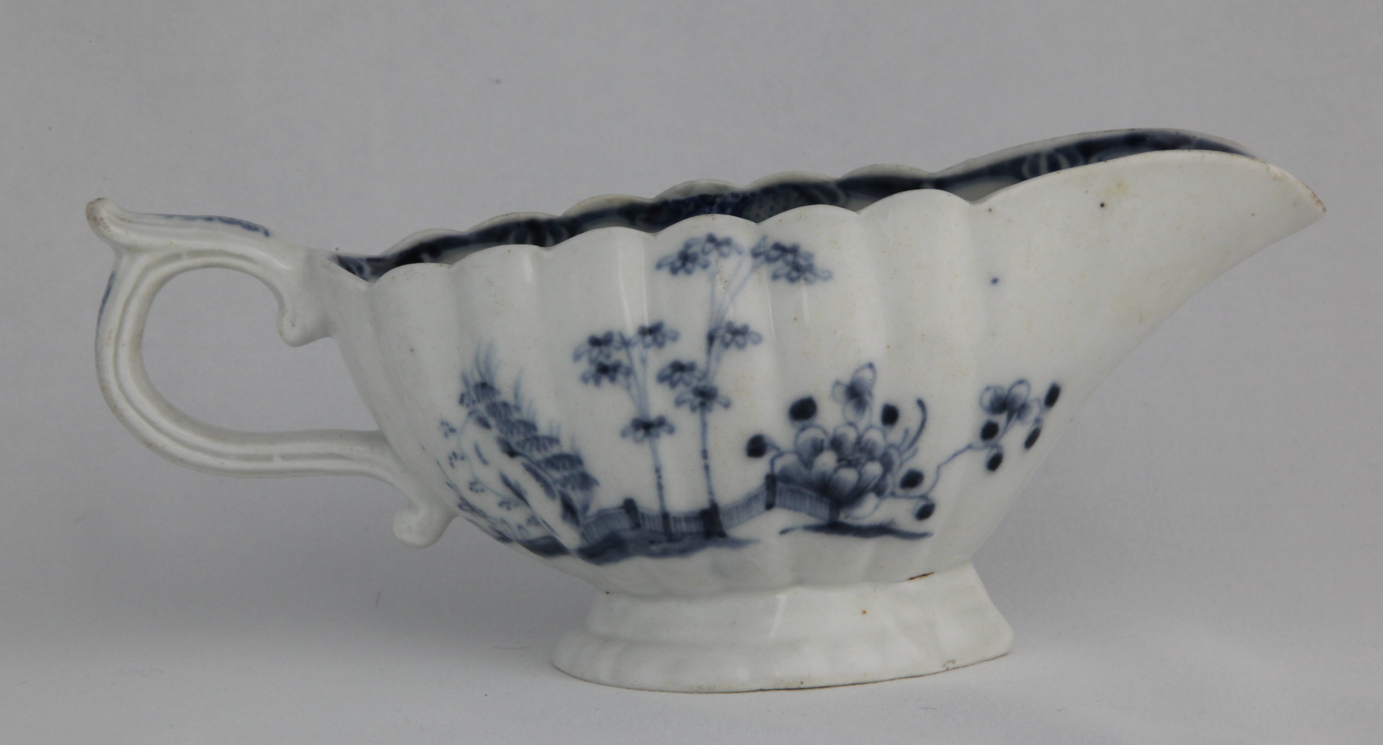 Sauceboat, from the Blue/white service, decorated by No mark, A Plymouth (William Cookworthy) sauceboat, circa 1768-70. Of fluted or gadrooned form, painted in blue with a version of the 'Plantation' pattern, bamboo and peony growing by rockwork and a fence, a scroll border inside the rim, 20.8cm long, tin or '24' mark — Plymouth & Bristol porcelain piece 1505