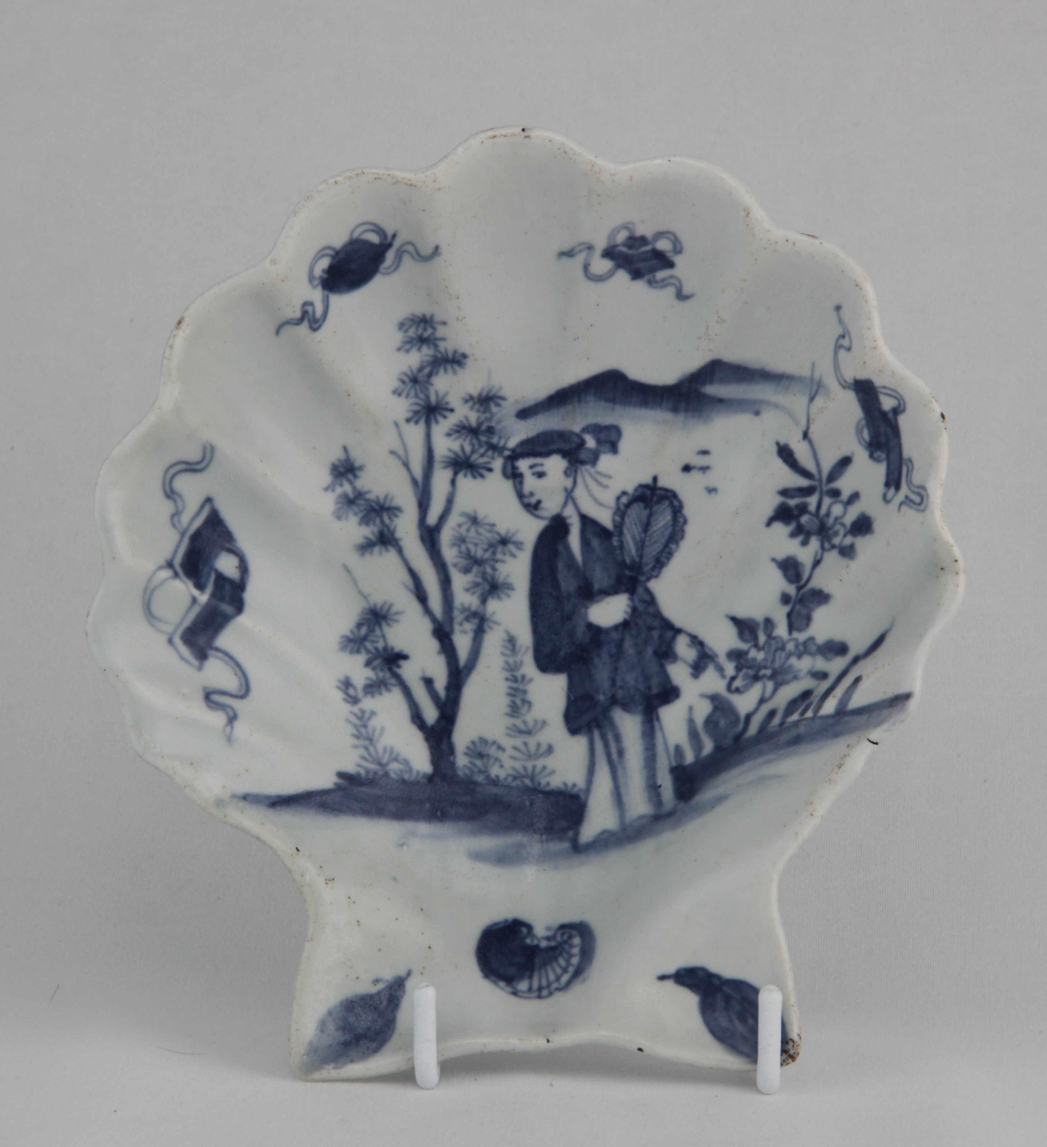 An exceptional Lund's Bristol large scallop shell dish, circa 1749, sharply moulded in the shape of a single scallop shell, painted in blue with a 'Long Eliza' figure holding a fan, standing beside a pine tree and pointing to a flowering plant, four ribboned 'Precious Objects' inside the rim and a shell and two leaves at the base, the underside rim painted with plant tendrils, 14.5cm high