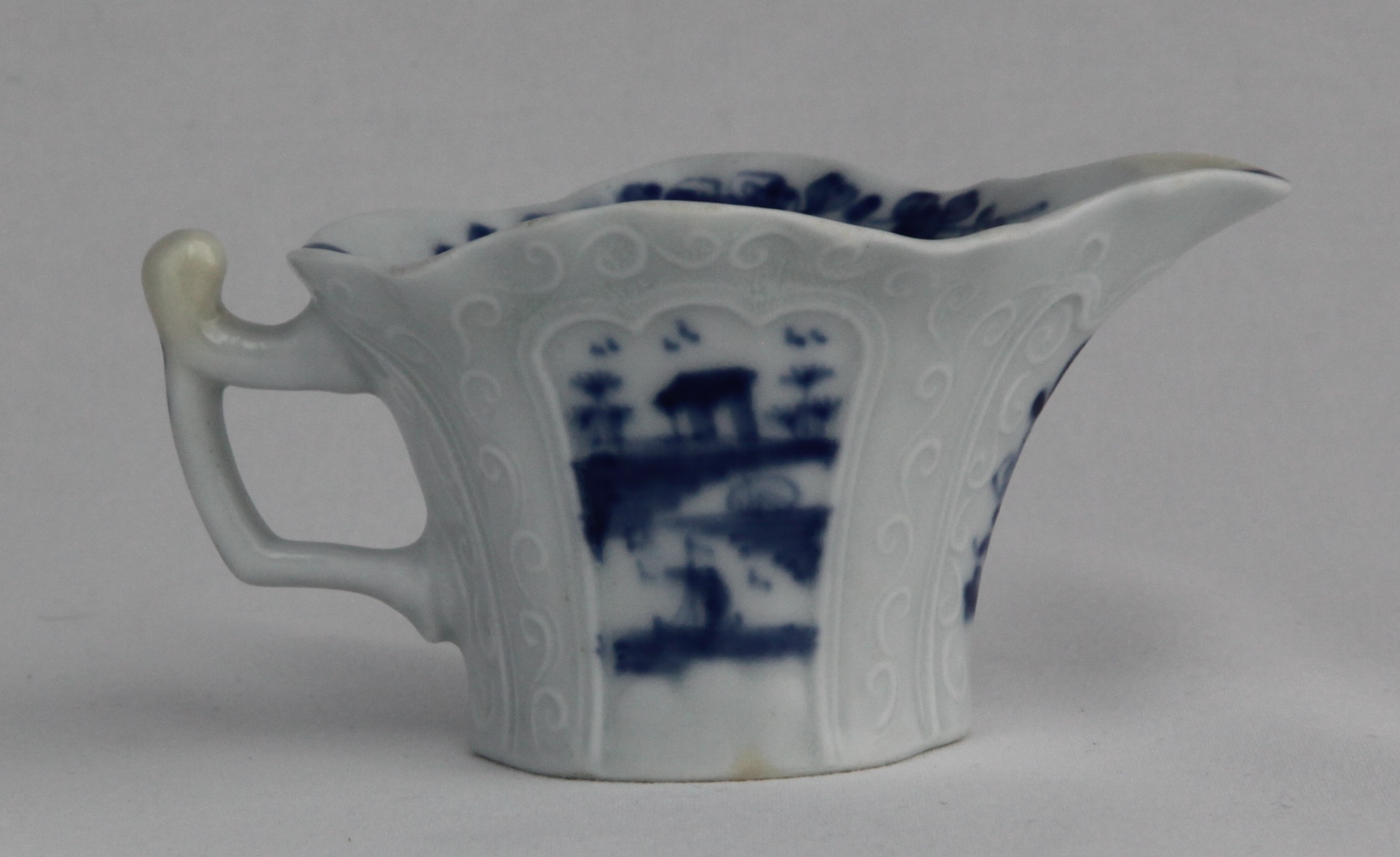 An important marked Lund's Bristol creamboat, circa 1749-50, of flared form with a gently undulating rim, the sides delicately moulded with 'double-rimmed' cartouches with tri-lobed tops flanked by chains of simple whorls, painted in blue, the front with a standing Chinese figure holding a flower, the reverse with a sampan, a hut and a 'floating rock', a flower sprig under the spout, shells, flowers and leaves around the interior rim, a further floating rock painted in the base, 10.7cm long, moulded mark in relief 'Bristol' to the base close to the handle