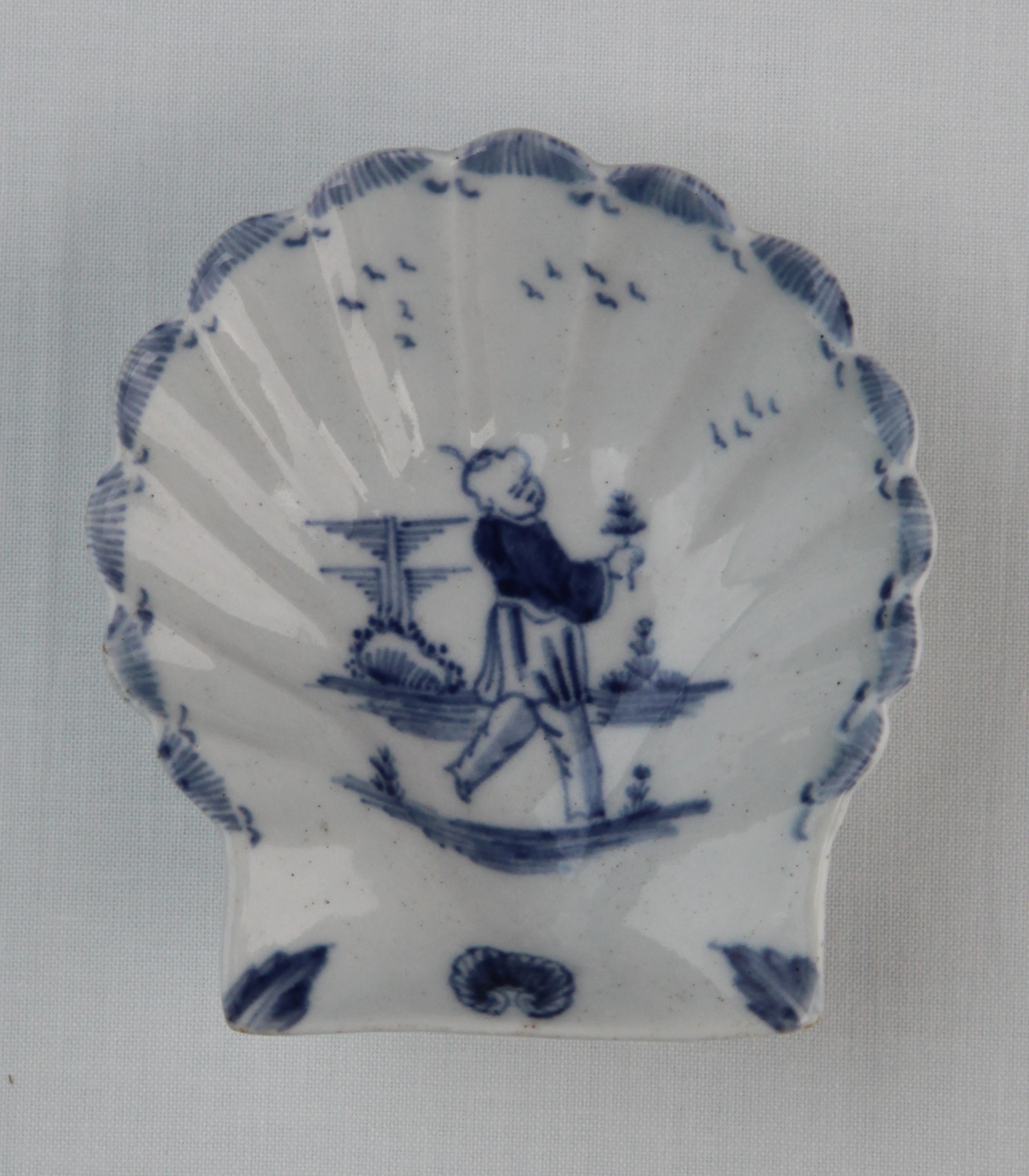 A Lund's Bristol scallop shell pickle dish, circa 1751, of small size, the underside moulded with chevrons around the footrim, painted in blue with a Chinese boy in a walking attitude, holding a flower, pylon foliage on a striped rock behind him and flights of birds above, the rim with close-packed horizontal lines, shell and leaf motifs at the base, 8.4cm high