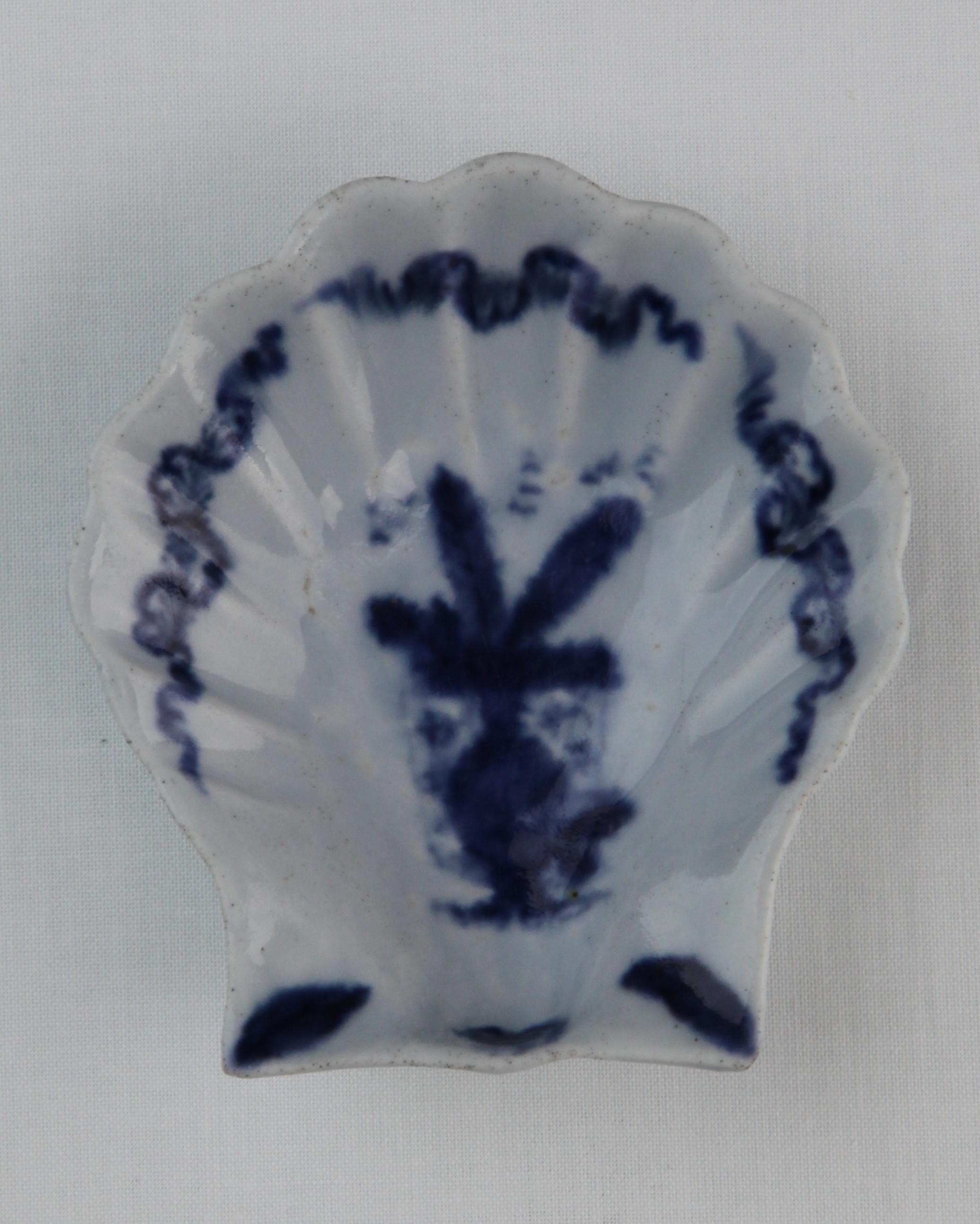 A Lund's Bristol scallop shell pickle dish, circa 1748-50, painted in blue with a Chinese vase containing two feathers in front of a partly unrolled scroll, the border with three long feathers entwined with ribbons, a shell flanked by two leaves at the base, the underside with two leafy branches, 9cm long