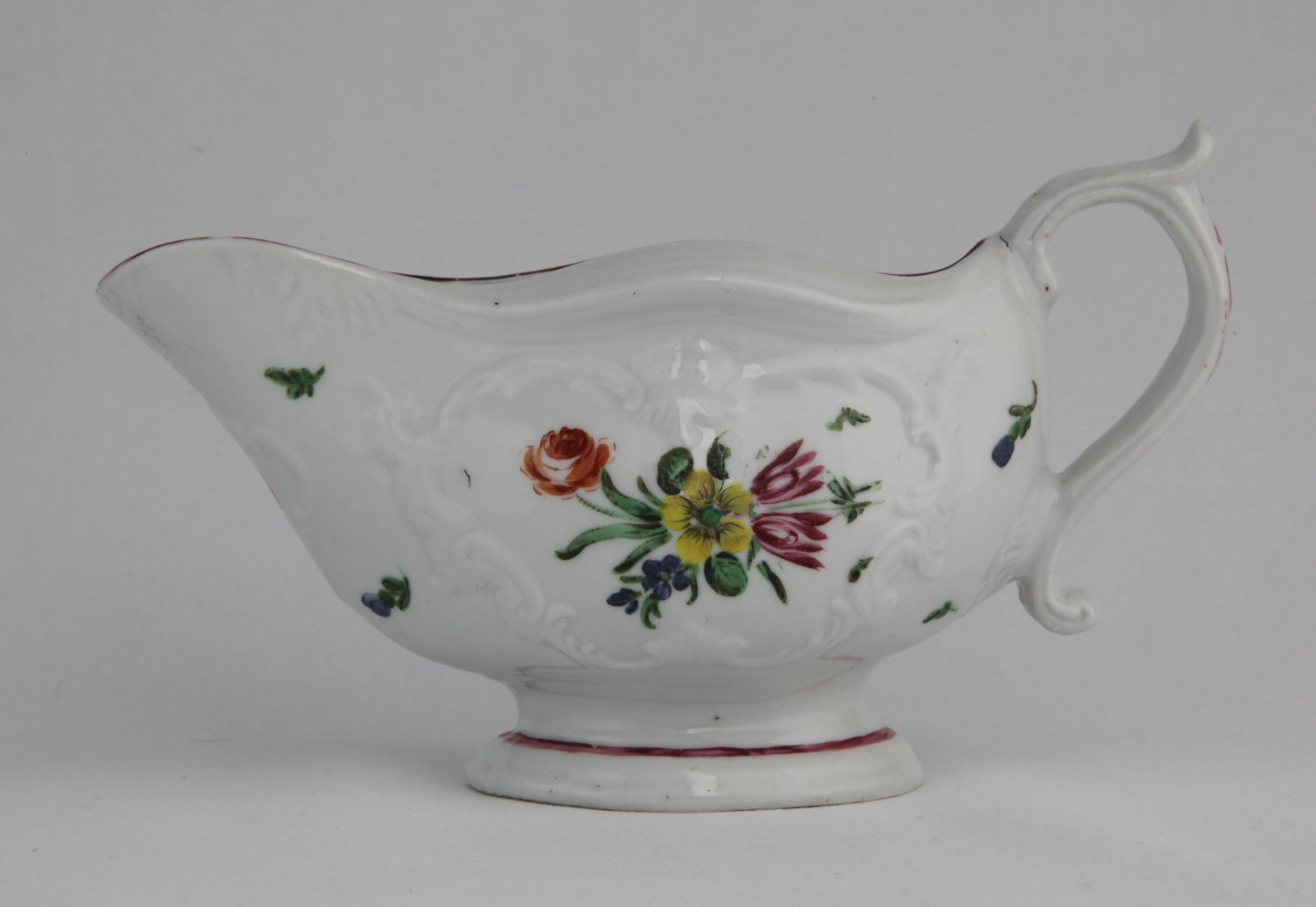A Bristol sauceboat, with wavy rim, scroll-framed moulded panel and scroll handle with thumbrest, painted in enamels with a colourful bouquet of flowers and scattered leaves, red rim line and red line around foot. X11. mark