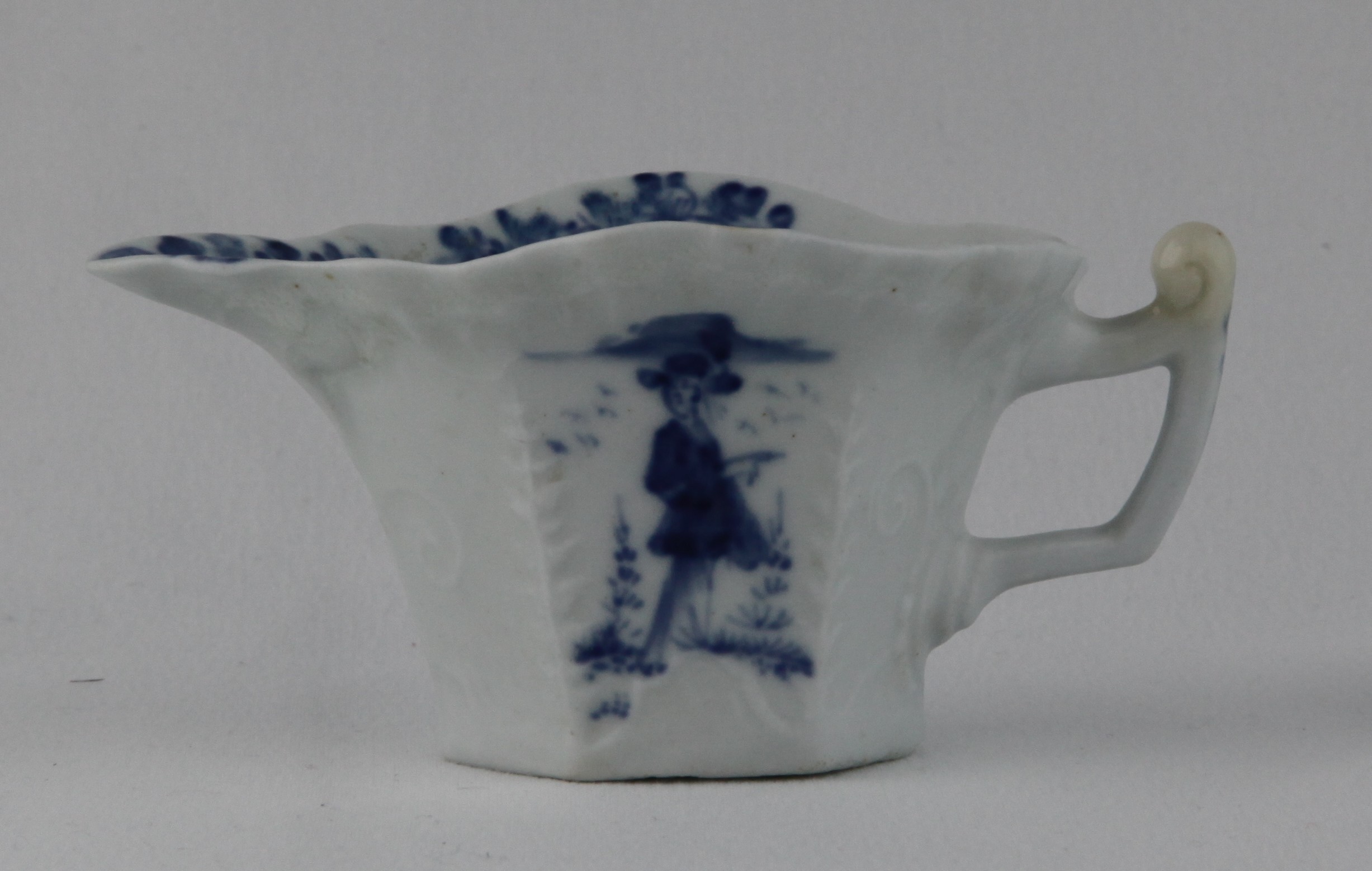 A rare Lund's Bristol creamboat, circa 1750 of flared hexagonal form with a shaped rim, the angular handle with an upturned thumbrest, delicately moulded with a geranium leaf below the lip, a feuilles-de-choux border below the rim, and scrolls around the base and handle, painted in blue on both sides with a 'Long Eliza' figure flanked by trees and birds, flower sprays to the interior, 11.1cm long