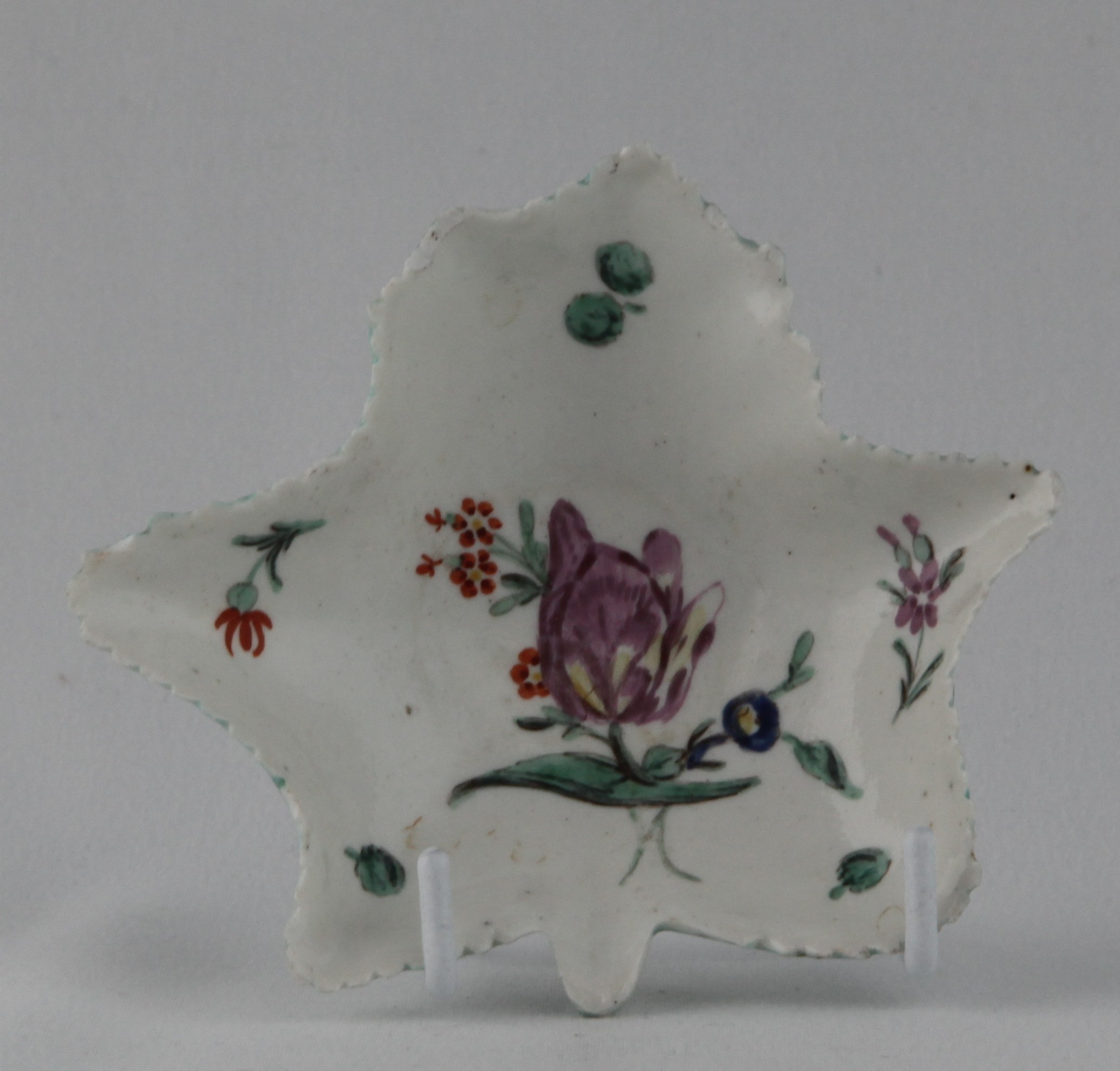 A Bristol leaf shaped pickle dish, boldly painted in coloured enamels with a purple tulip and other flowers, and with scattered sprigs, 4 1/8" wide, circa 1772, pseudo Meissen underglaze blue crossed swords mark and numeral 17 in blue