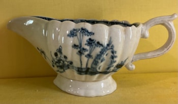 A Plymouth fluted oval sauceboat, with notched scroll handle, painted in underglaze blue in Chinese style with bamboo, peony and rockwork, in fenced gardens, the interior painted with a flower, within a diaper panelled scroll border, 7 3/4" long, circa 1770, blue painted tin and X marks