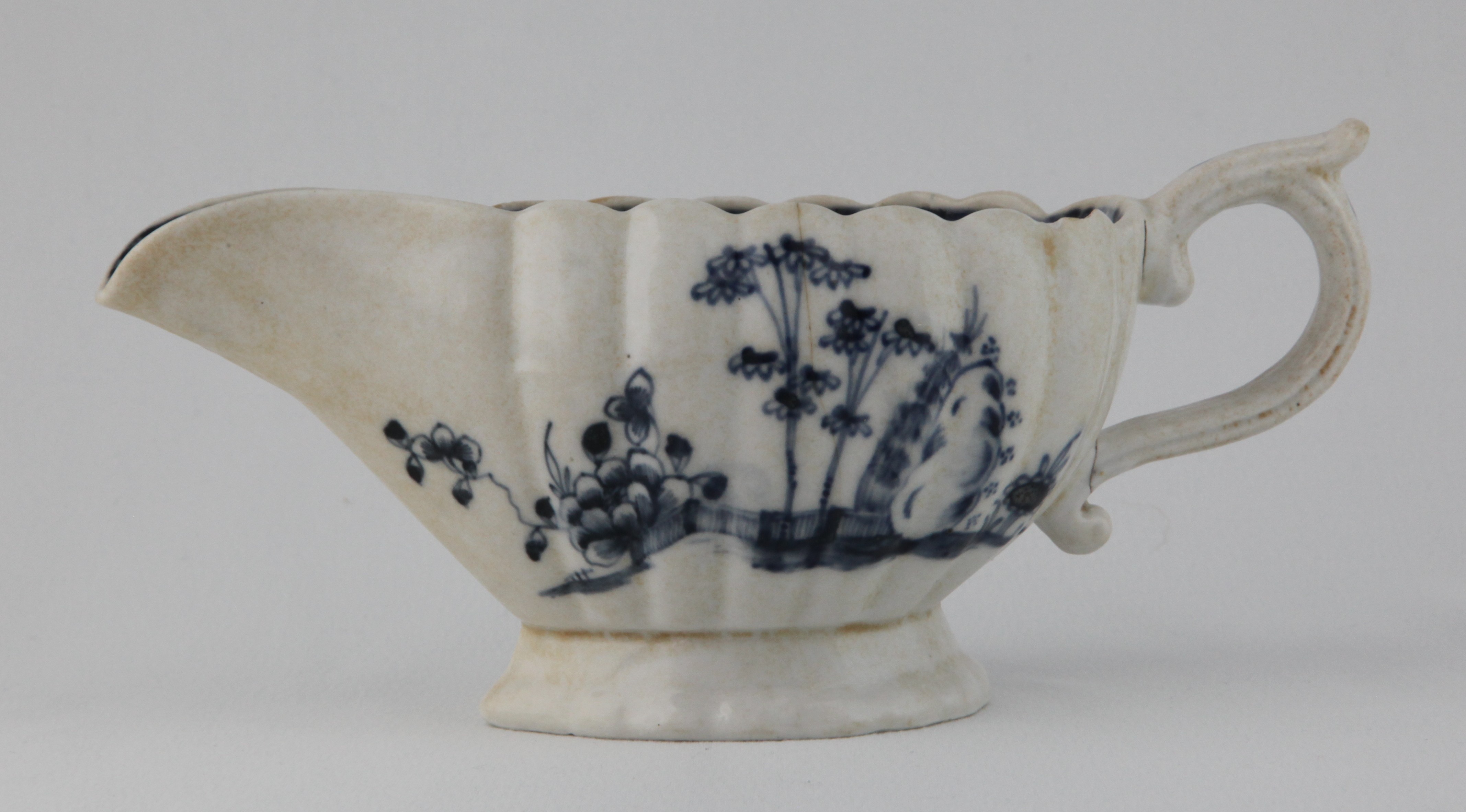 A Plymouth fluted oval sauceboat, with notched scroll handle, painted in underglaze blue in Chinese style with bamboo, peony and rockwork, in fenced gardens, the interior painted with a flower, within a diaper panelled scroll border, 7 3/4" long, circa 1770, blue painted tin and X marks