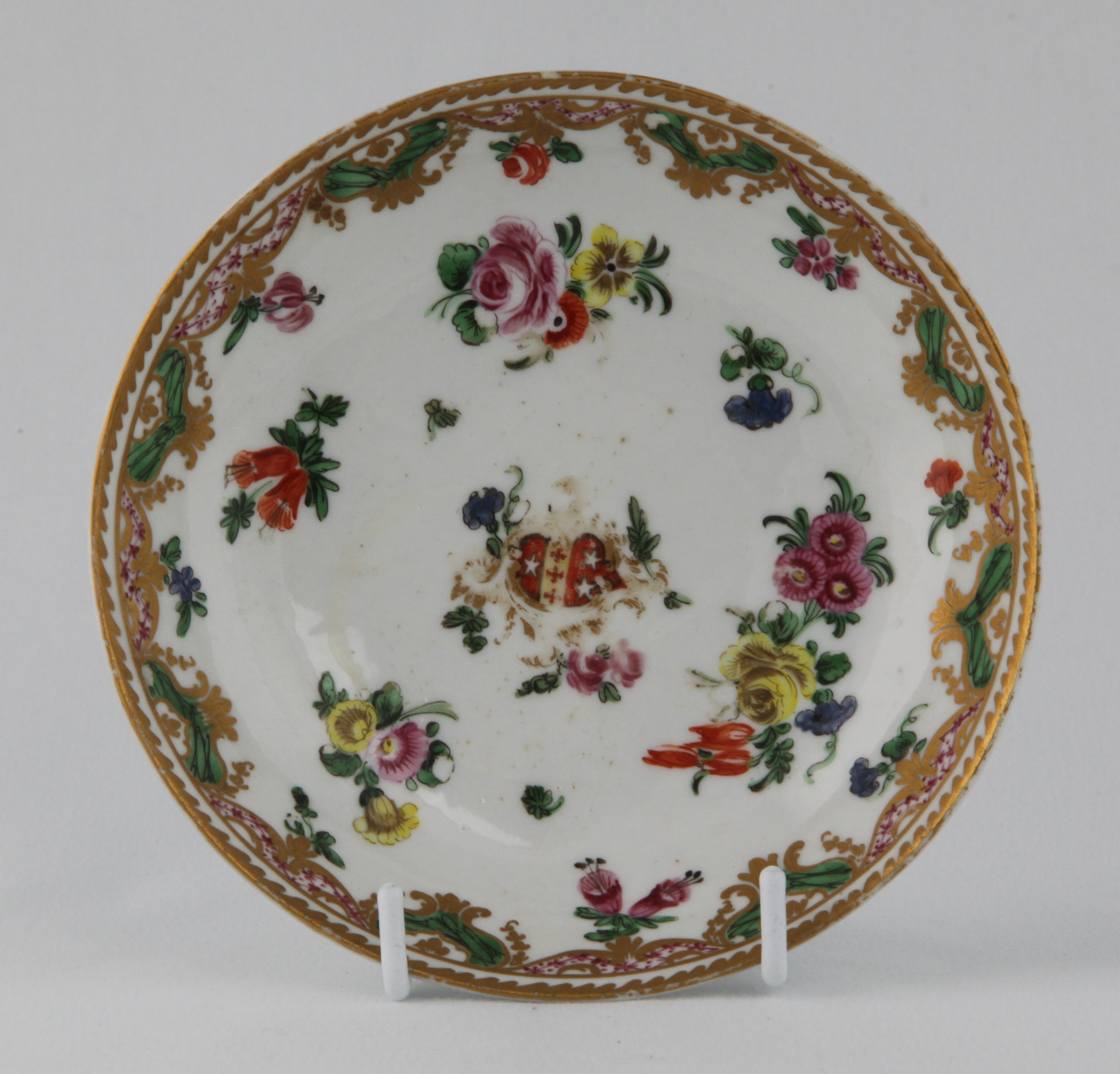 A Bristol tea saucer, from the Ludlow of Campden Service, painted with the arms of Daniel Ludlow of Campden, within a gilt scroll cartouche, and with flower sprays, the borders painted with flowers, suspended from a puce, green and gilt diaper panelled band, angular gilt dentil rims, circa 1775, no mark