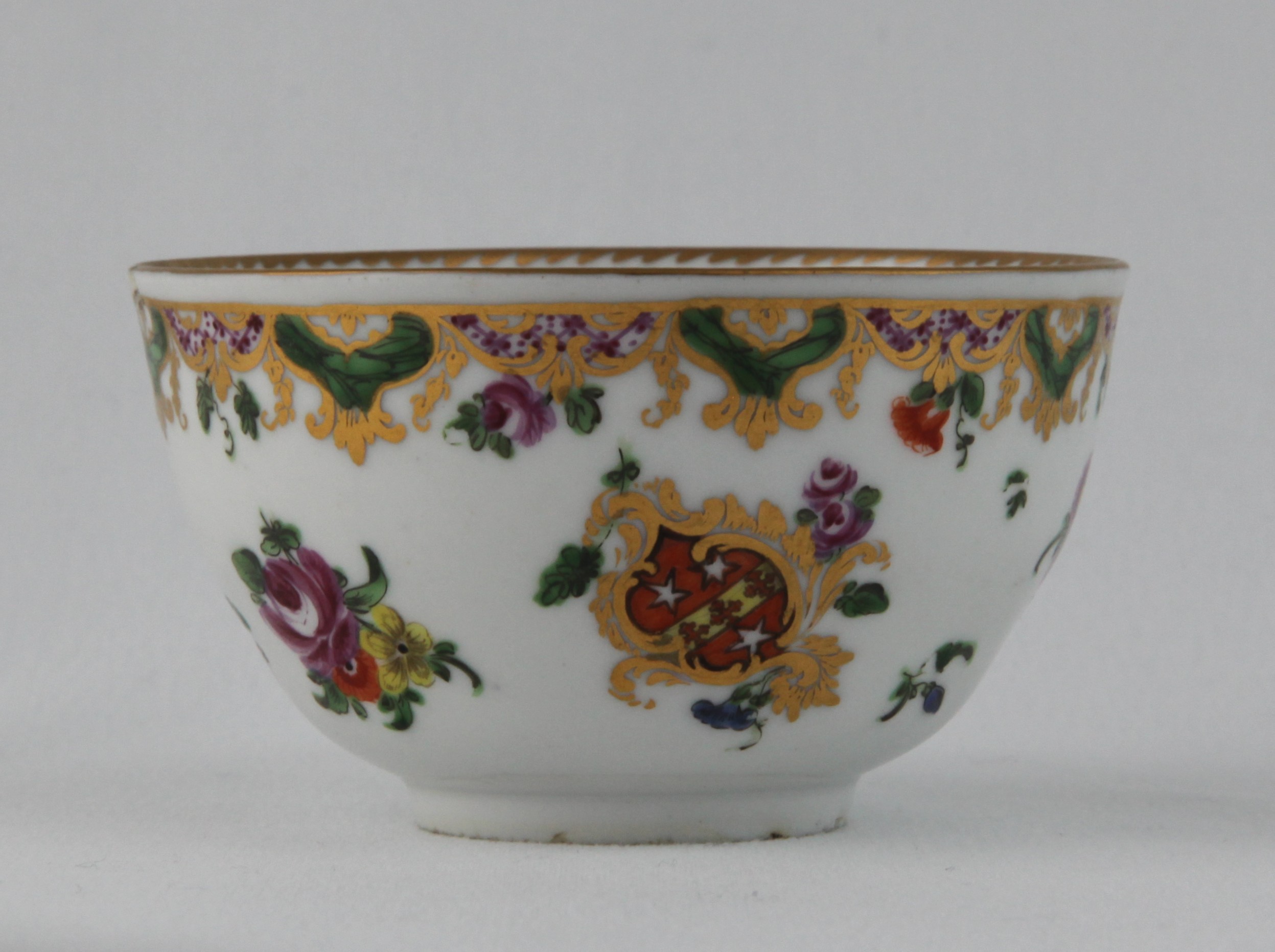 A Bristol teacup from the Ludlow of Campden Service, with entwined loop handle,  painted with the arms of Daniel Ludlow of Campden, within a gilt scroll cartouche, and with flower sprays, the borders painted with flowers, suspended from a puce, green and gilt diaper panelled band, angular gilt dentil rims, circa 1775, no mark