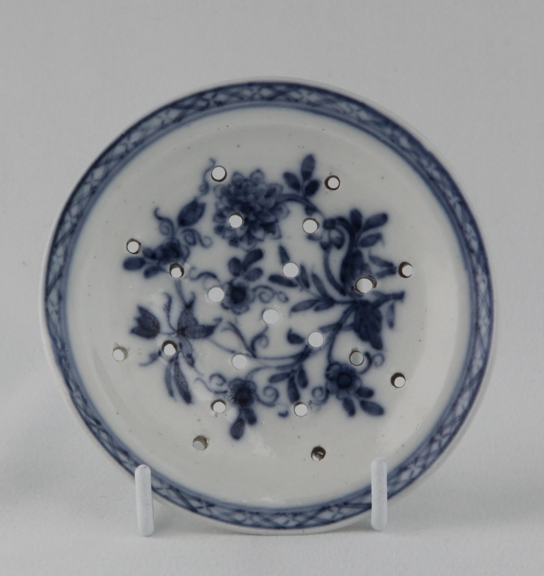 A very rare Bristol pierced round drainer, painted in underglaze blue with a stem of scrolling flowers and leaves, within a diaper border, 4" diameter, circa 1772, blue painted X mark