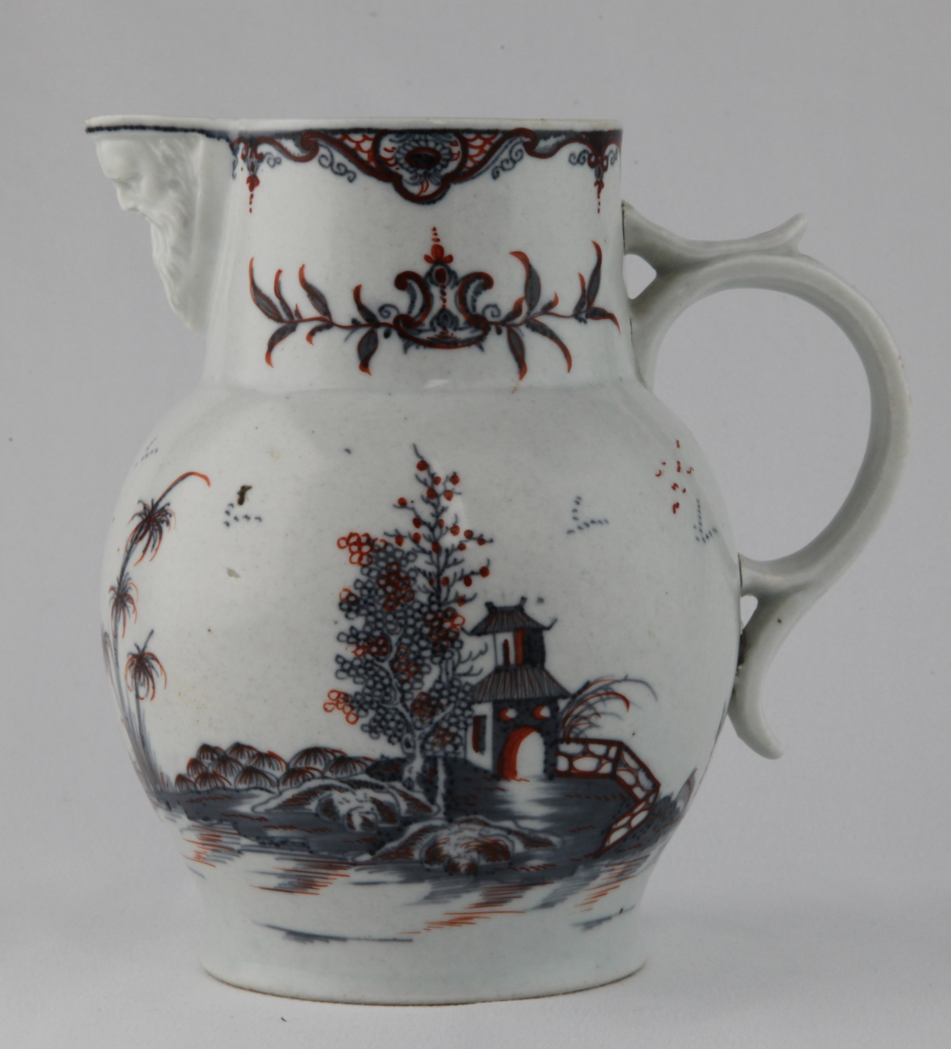 A Plymouth ovoid jug, with straight neck and male mask spout, and 'C' scroll handle, painted in underglaze blue in Chinese style with buildings in a river landscape, and picked out in iron red enamel, beneath trailing flower sprays and a flower and diaper panelled border, 6 1/2" high, circa 1770.