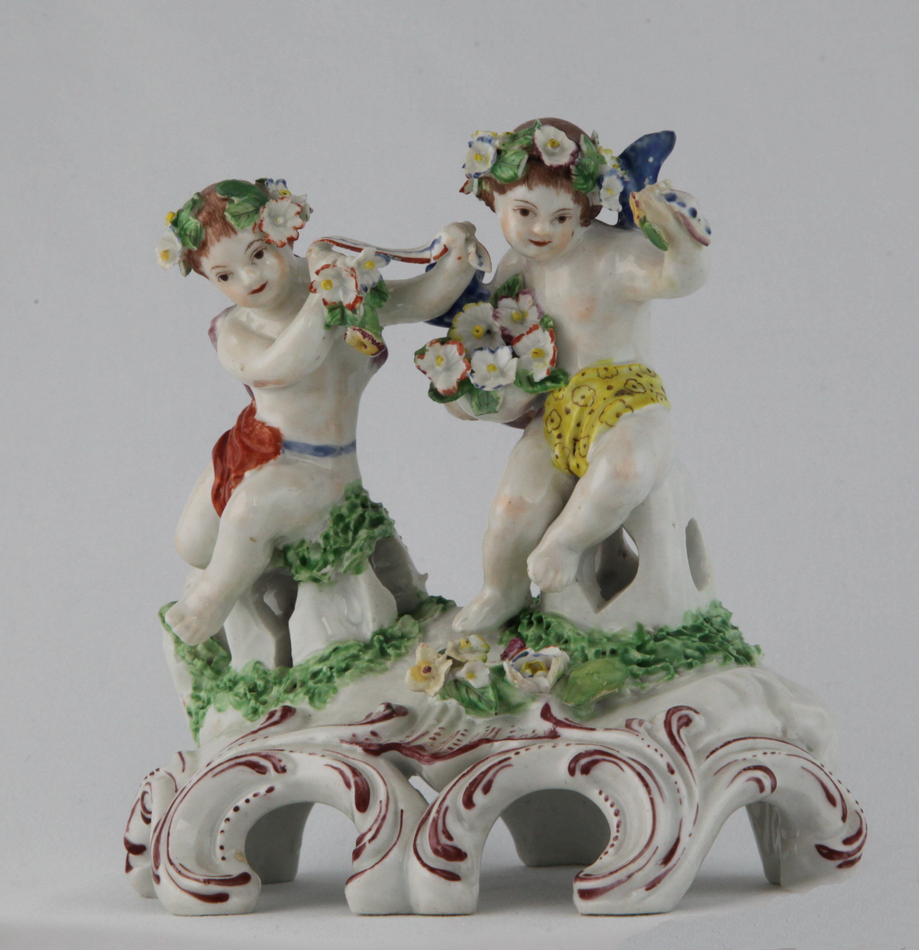 A Plymouth group of two garlanded putti, one holding a tub of flowers, and the other with a ribbon, decorated in coloured enamels, the scroll moulded mound base applied with flowers and leaves, and picked out in puce, 5 1/2" high, circa 1770, no mark
