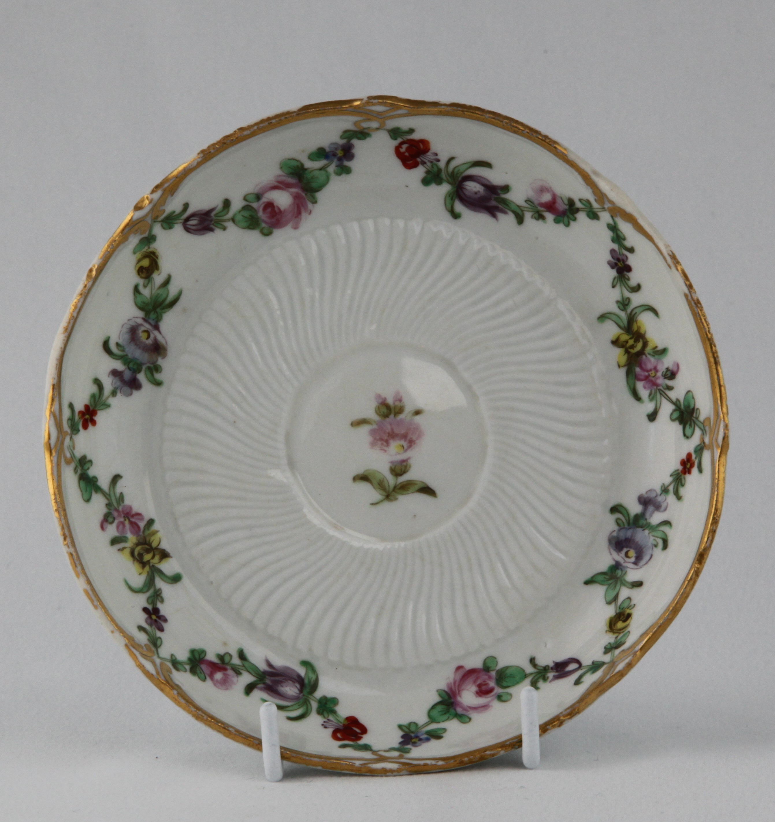 A Bristol coffee saucer painted in coloured enamels with swags of flowers and leaves, suspended from gilt loops, on a part spirally fluted ground, within gilt 'C' scroll borders, circa 1774-75, blue painted X marks and numeral 1