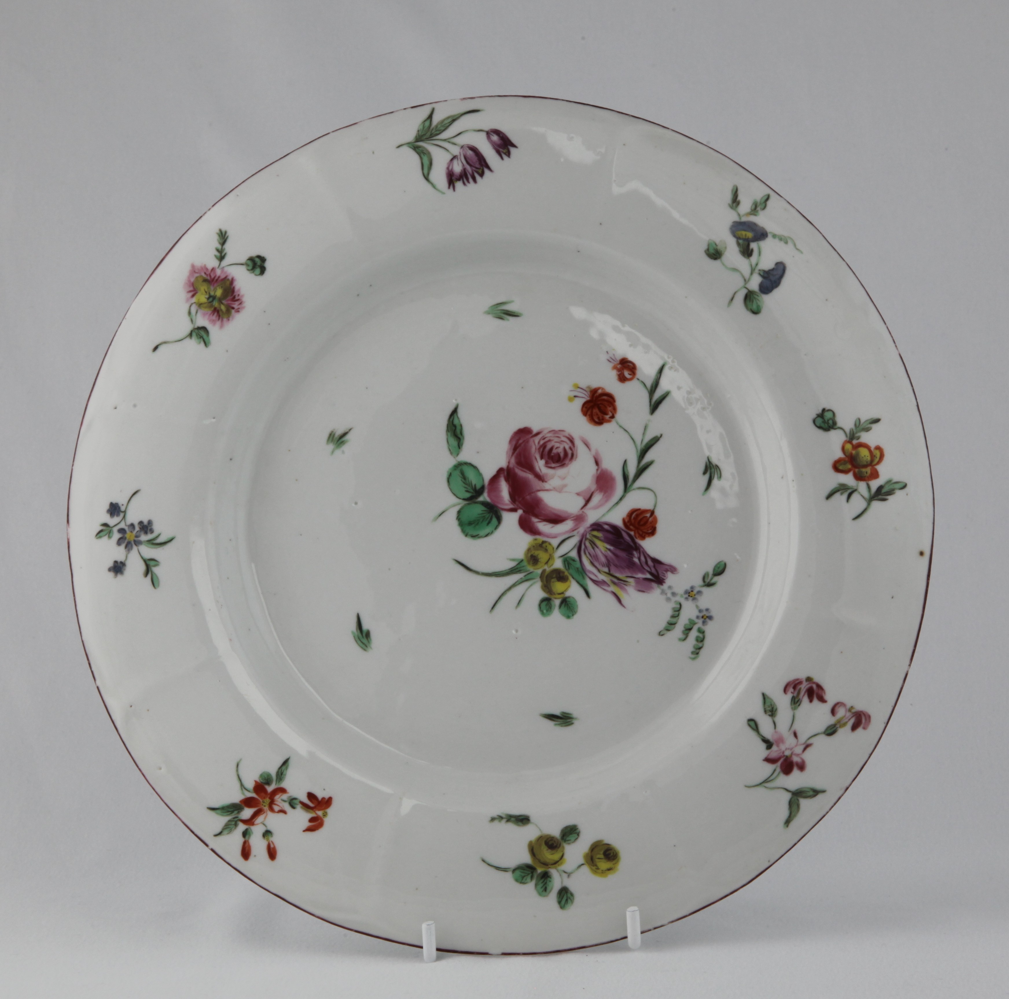 An unusual Bristol round plate, with slightly fluted border, painted in coloured enamels with a spray of flowers and leaves, and scattered flowers, brown line rim, 9 1/4" diameter, circa 1772, blue painted X mark and numeral 17