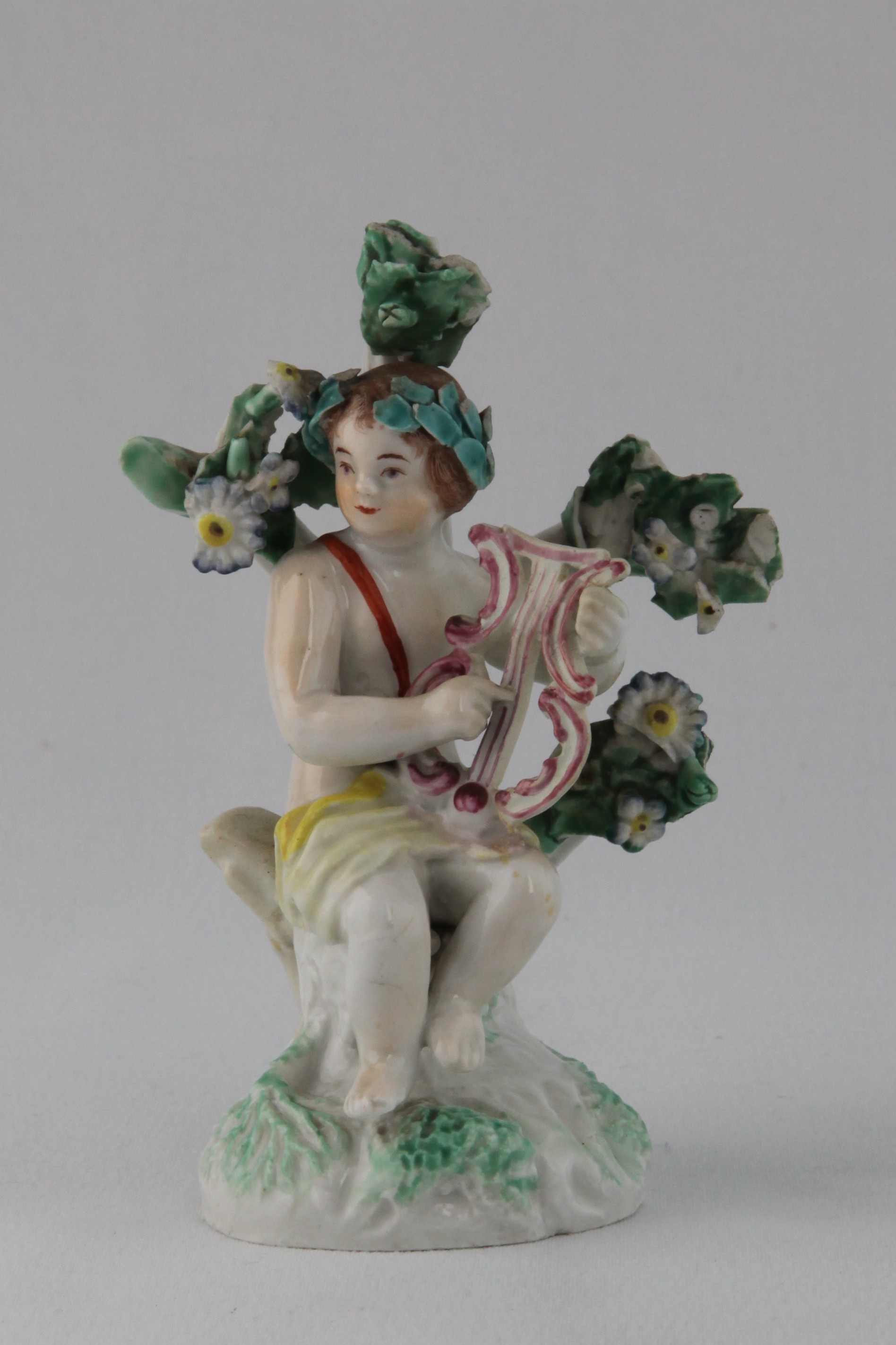 The lyre player of a rare pair of Plymouth small figures of garlanded putti, one playing a lyre and the other a lute, picked out in puce, brown and yellow, and seated before floral bocage, on leaf moulded mound base, 4" high, circa 1770, no marks