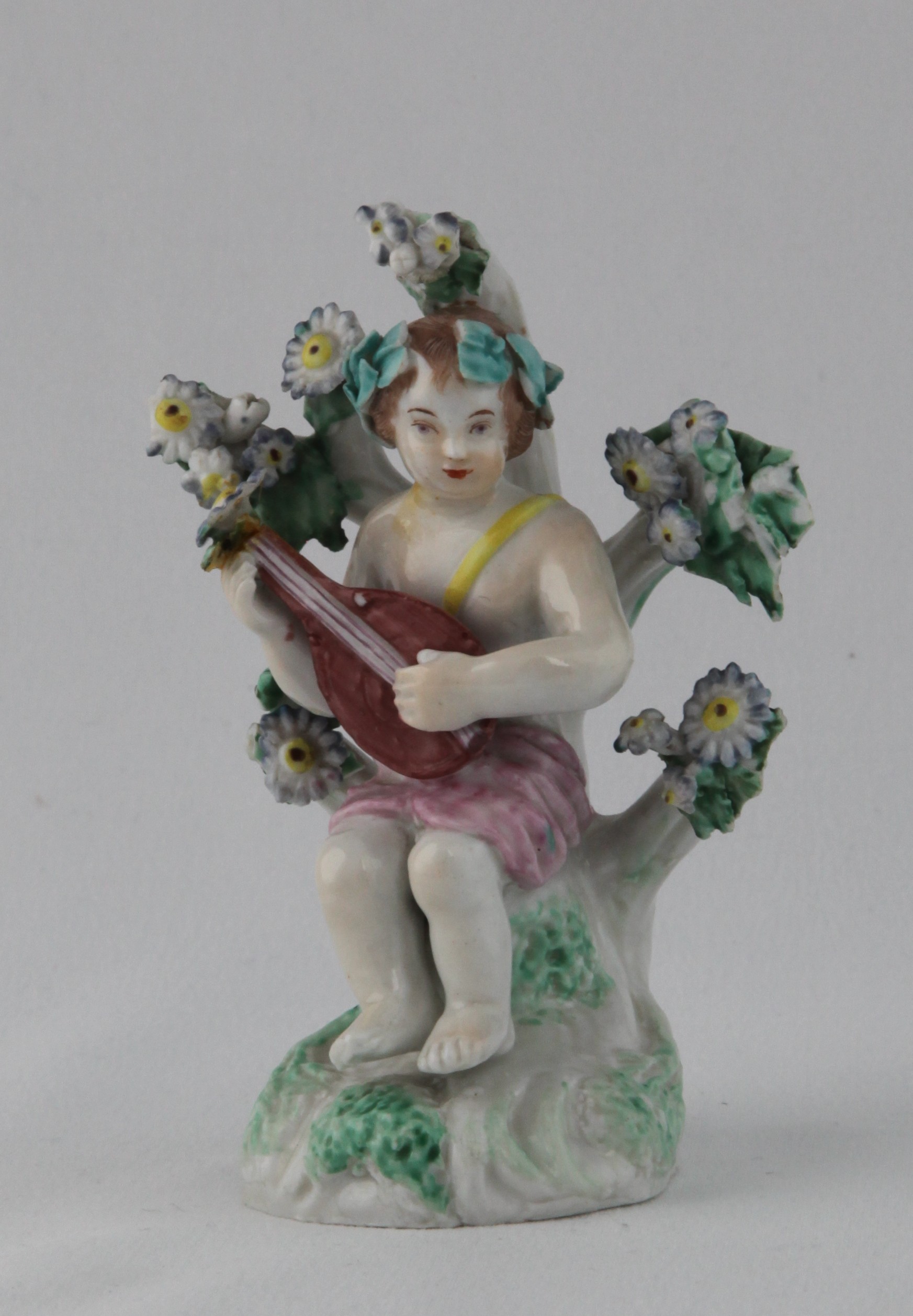 The lute player of a rare pair of Plymouth small figures of garlanded putti, one playing a lyre and the other a lute, picked out in puce, brown and yellow, and seated before floral bocage, on leaf moulded mound base, 4" high, circa 1770, no marks
