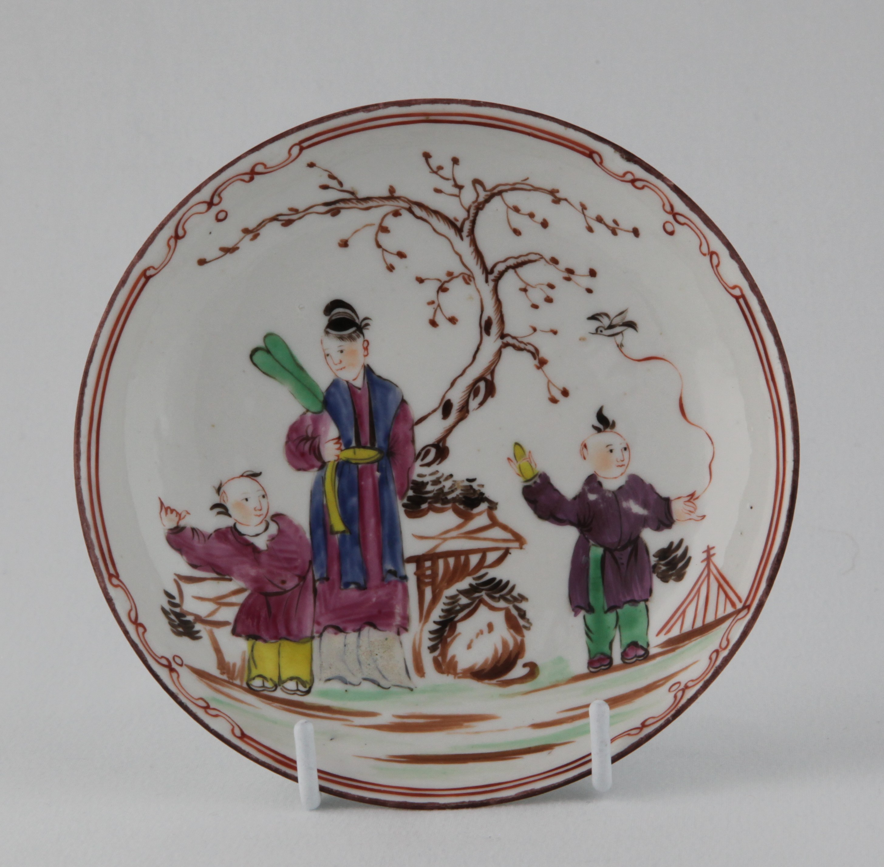 A Bristol saucer, painted in coloured enamels in Chinese Export style with a mother and two children before a tree, in a fenced garden, within an iron red line and scroll border, 5" diameter, circa 1772, blue painted B mark and numeral 6