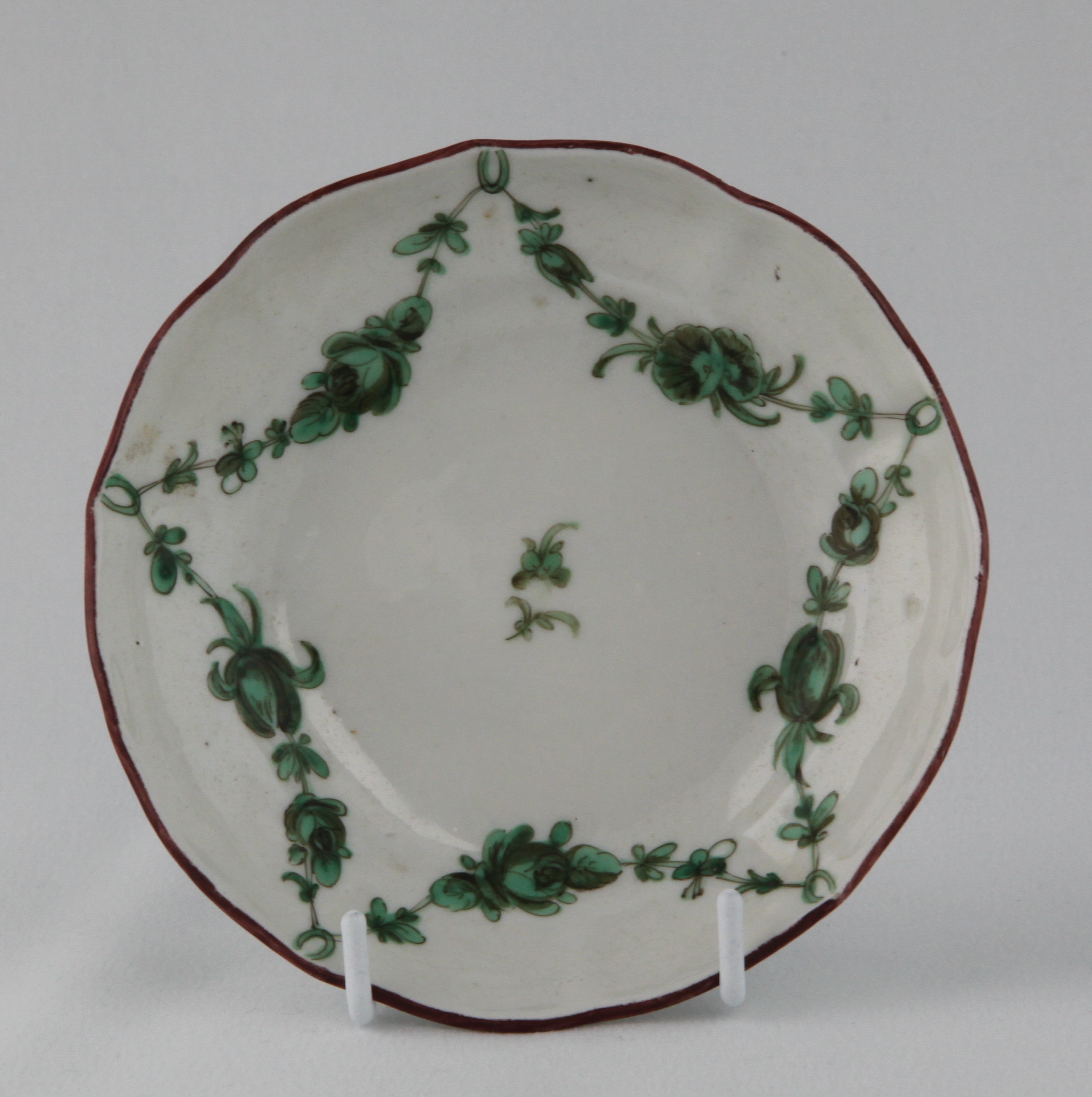 A Bristol small ogee shaped saucer, painted in green and black with floral garlands suspended from rings, within brown line rims, circa 1775, blue painted X marks and numeral 1.