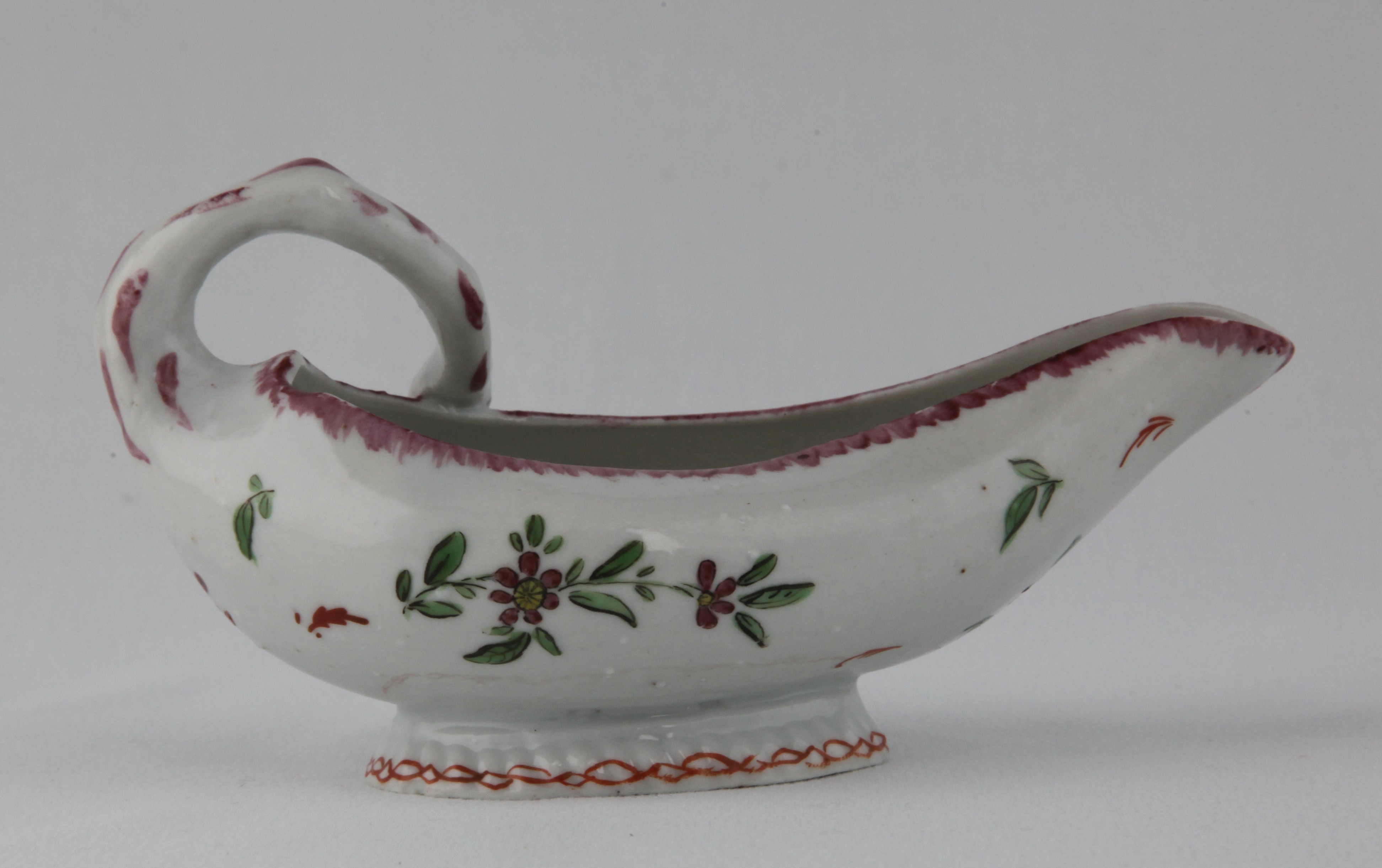 One of a pair of Bristol small leaf shaped sauceboats, each with stalk loop handle, painted in coloured enamels with flowers and leaves, beneath a puce feuille de choux border, each foot with an iron red interlinked band, 5 1/2" long, circa 1772, no marks