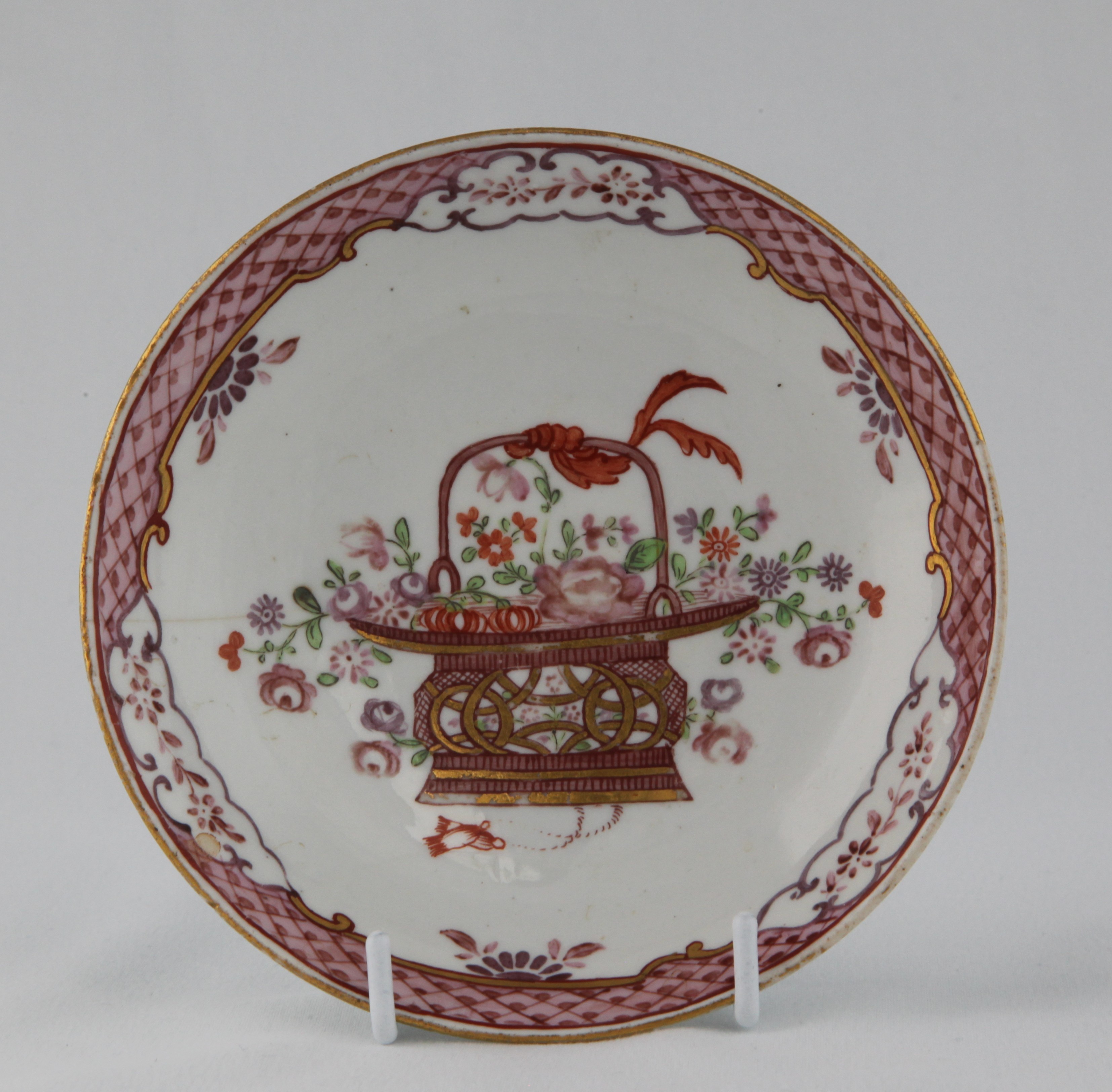 A Bristol saucer, finely painted in Chinese Export famille rose palette with a basket of flowers, within a flower panelled puce diaper border, 5 1/8" diameter, circa 1775, blue painted X mark and numeral 7.