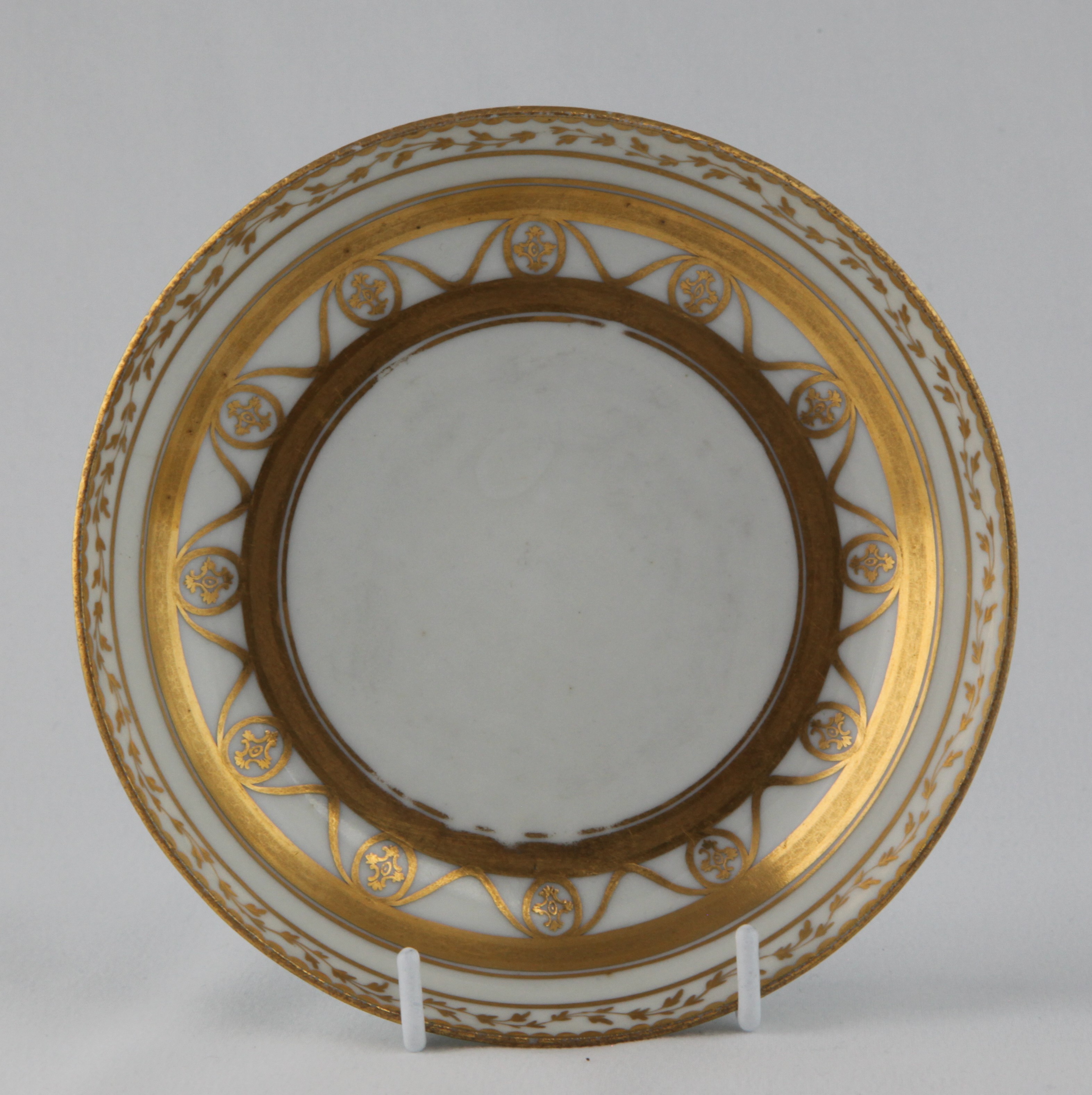 A Bristol saucer, unusually decorated in gilt with flower heads within oval panels, united by swags, within broad gilt bands, and bands of scrolling leaves, gilt dentil rims, circa 1775, blue painted X mark and numeral 4 in gilt