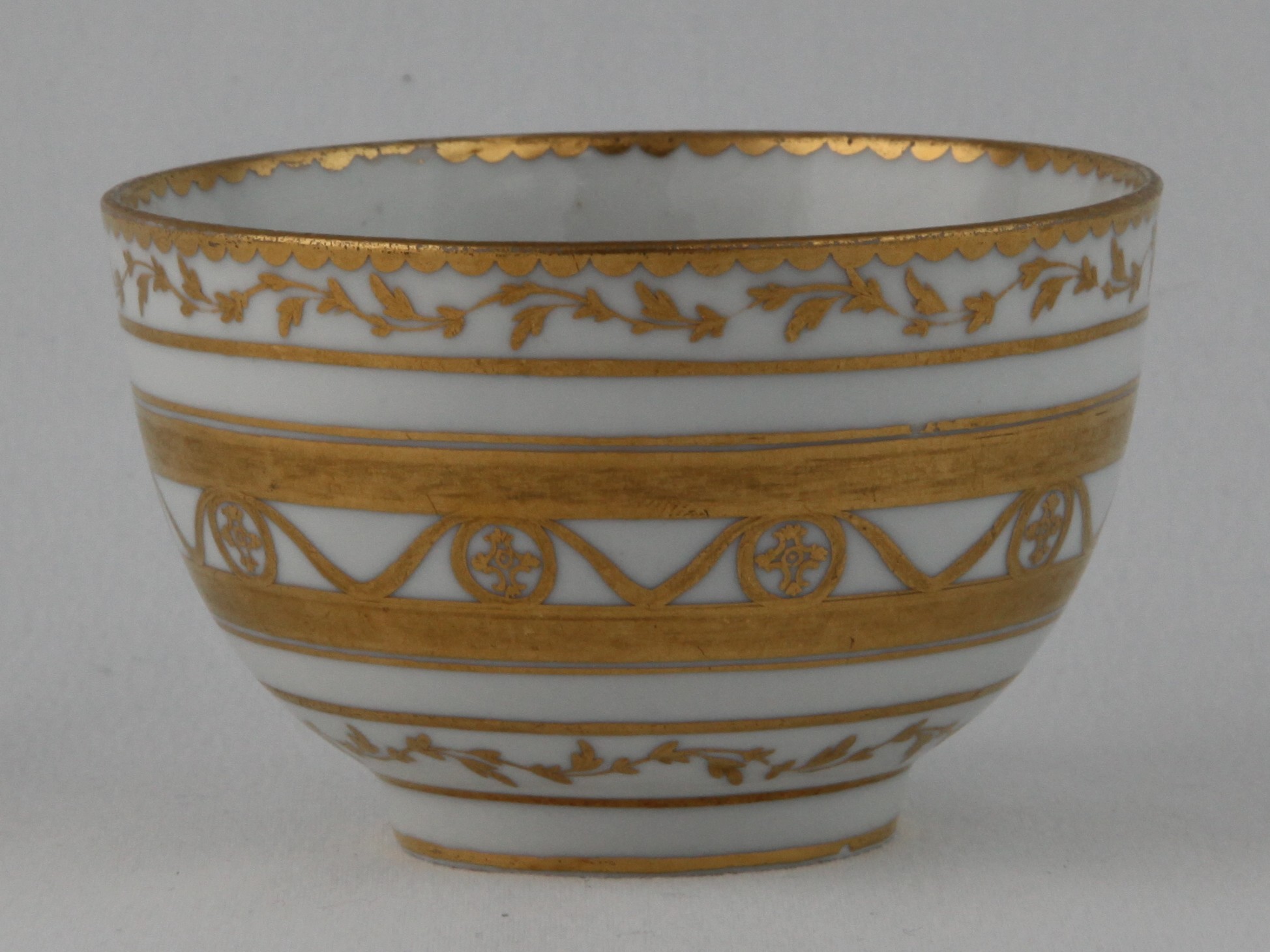 A Bristol tea bowl, unusually decorated in gilt with flower heads within oval panels, united by swags, within broad gilt bands, and bands of scrolling leaves, gilt dentil rims, circa 1775, blue painted X mark and numeral 4 in gilt