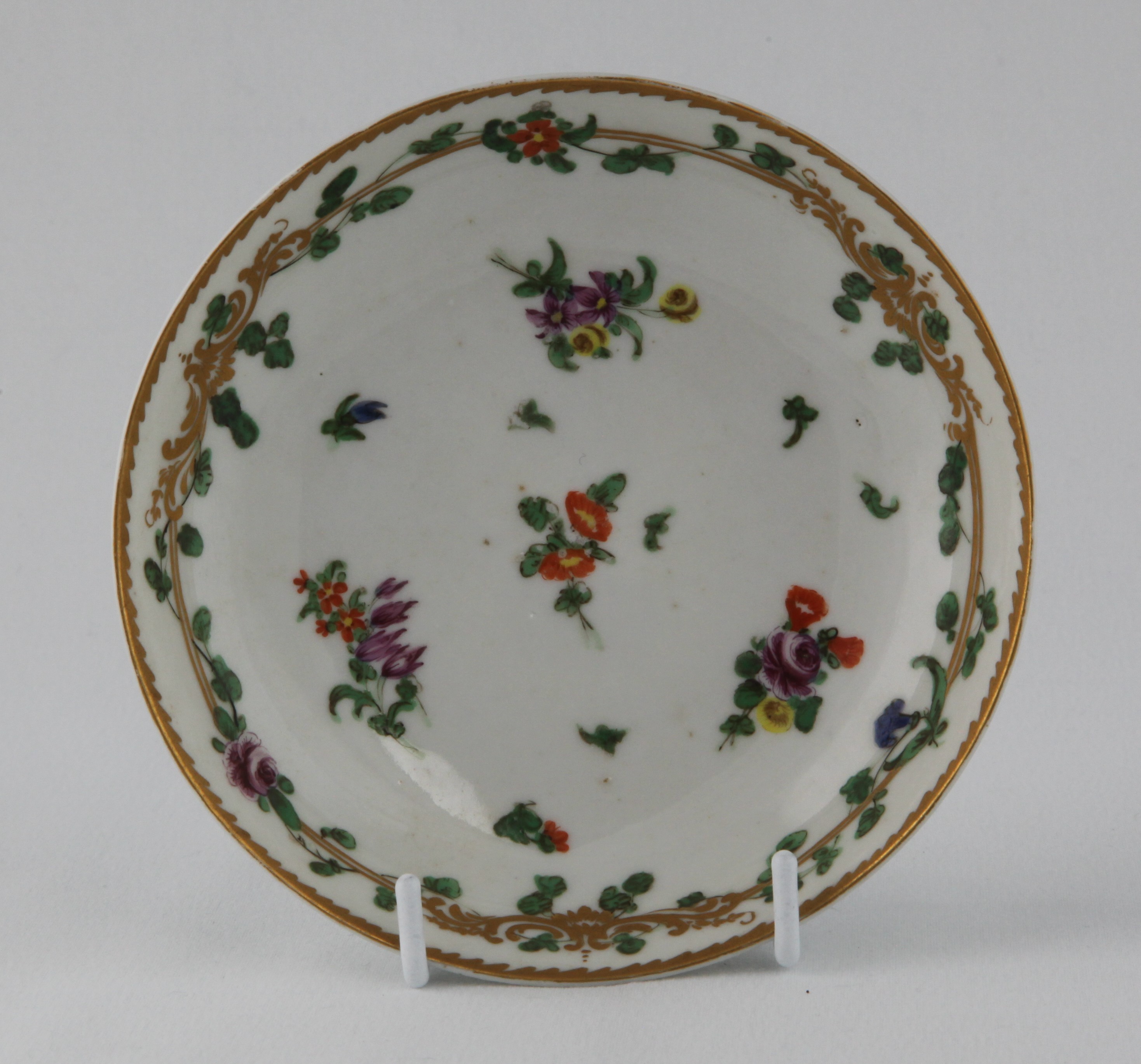 A Bristol ogee shaped saucer, painted in coloured enamels with sprays of flowers and leaves, and scattered flowers, within a gilt band entwined with flowers and leaves, angular gilt dentil rim, circa 1772, no marks