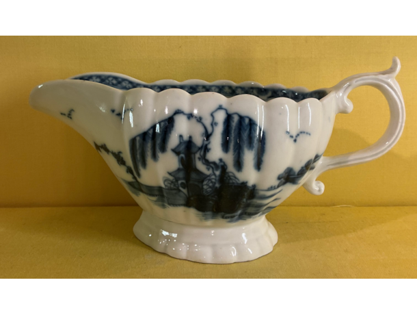 A Plymouth fluted oval sauceboat, the 'S' scroll handle with leaf moulded thumb piece, painted in underglaze blue in Chinese style with pavilions and willow trees in river landscapes, the interior painted with a flower, within a flower head panelled diaper border, 7 1/2" long, circa 1770, blue painted tin mark