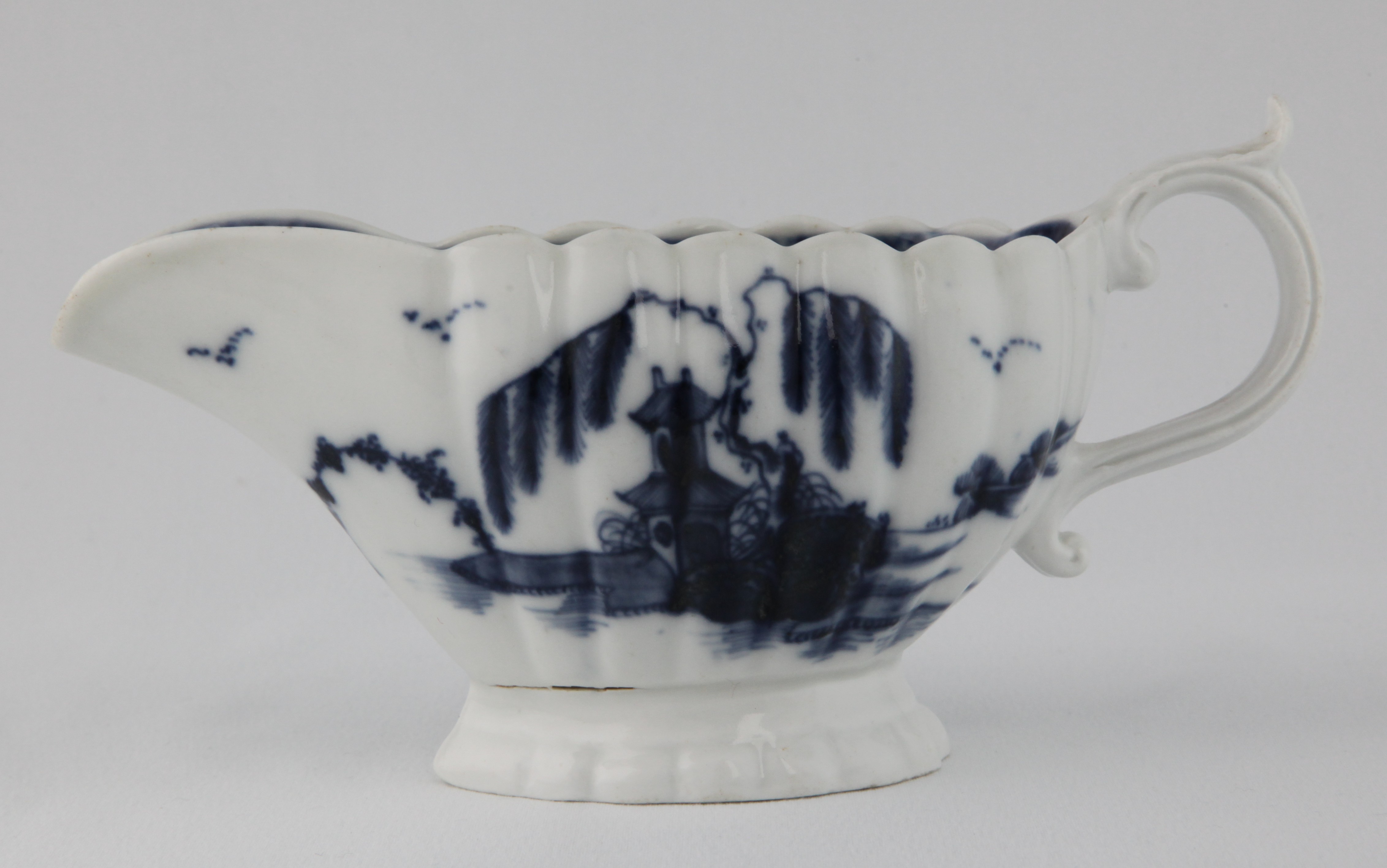 A Plymouth fluted oval sauceboat, the 'S' scroll handle with leaf moulded thumb piece, painted in underglaze blue in Chinese style with pavilions and willow trees in river landscapes, the interior painted with a flower, within a flower head panelled diaper border, 7 1/2" long, circa 1770, blue painted tin mark