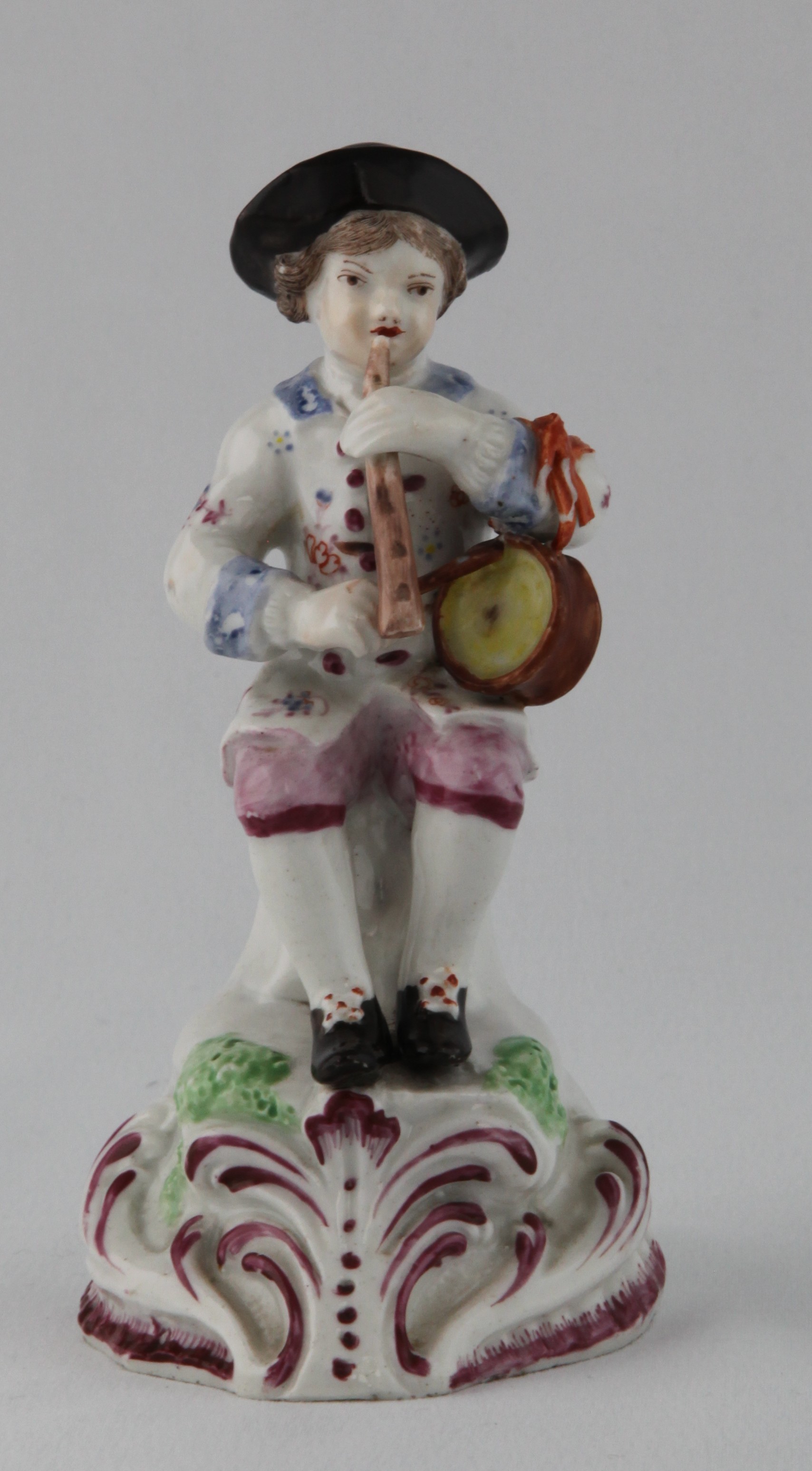 A Plymouth figure of a young boy seated on a tree stump, playing a pipe and a drum, decorated in coloured enamels, the high scroll moulded mound base picked out in puce, 5 3/4" high overall, circa 1770, no mark