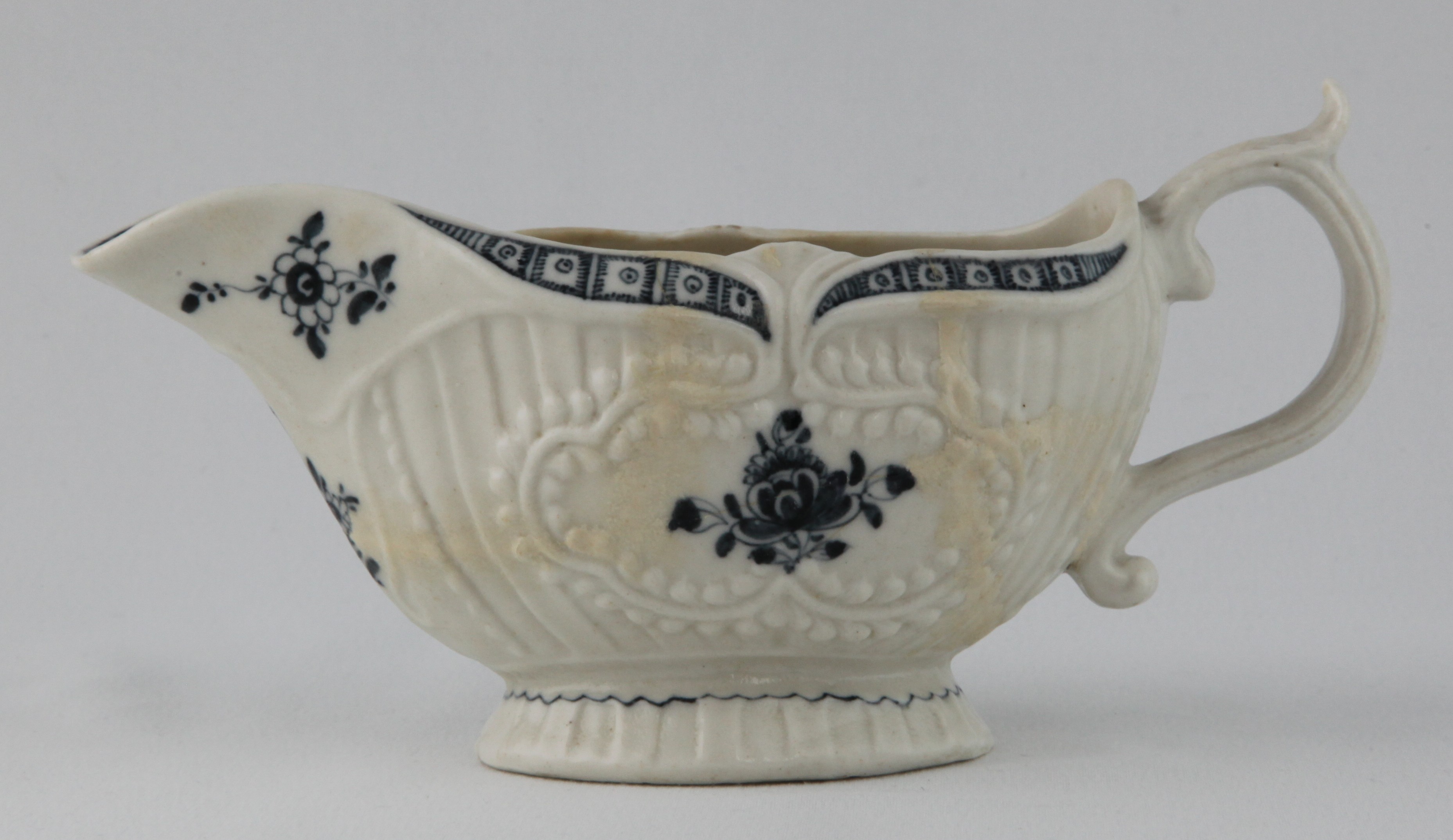 A Plymouth oval sauceboat, the 'S' loop handle with leaf scroll thumb piece, painted in underglaze blue with flower sprays, within beaded cartouches, on a strap fluted ground, the rim painted with a cell diaper band, 7 1/2" long, circa 1770, blue painted tin mark
