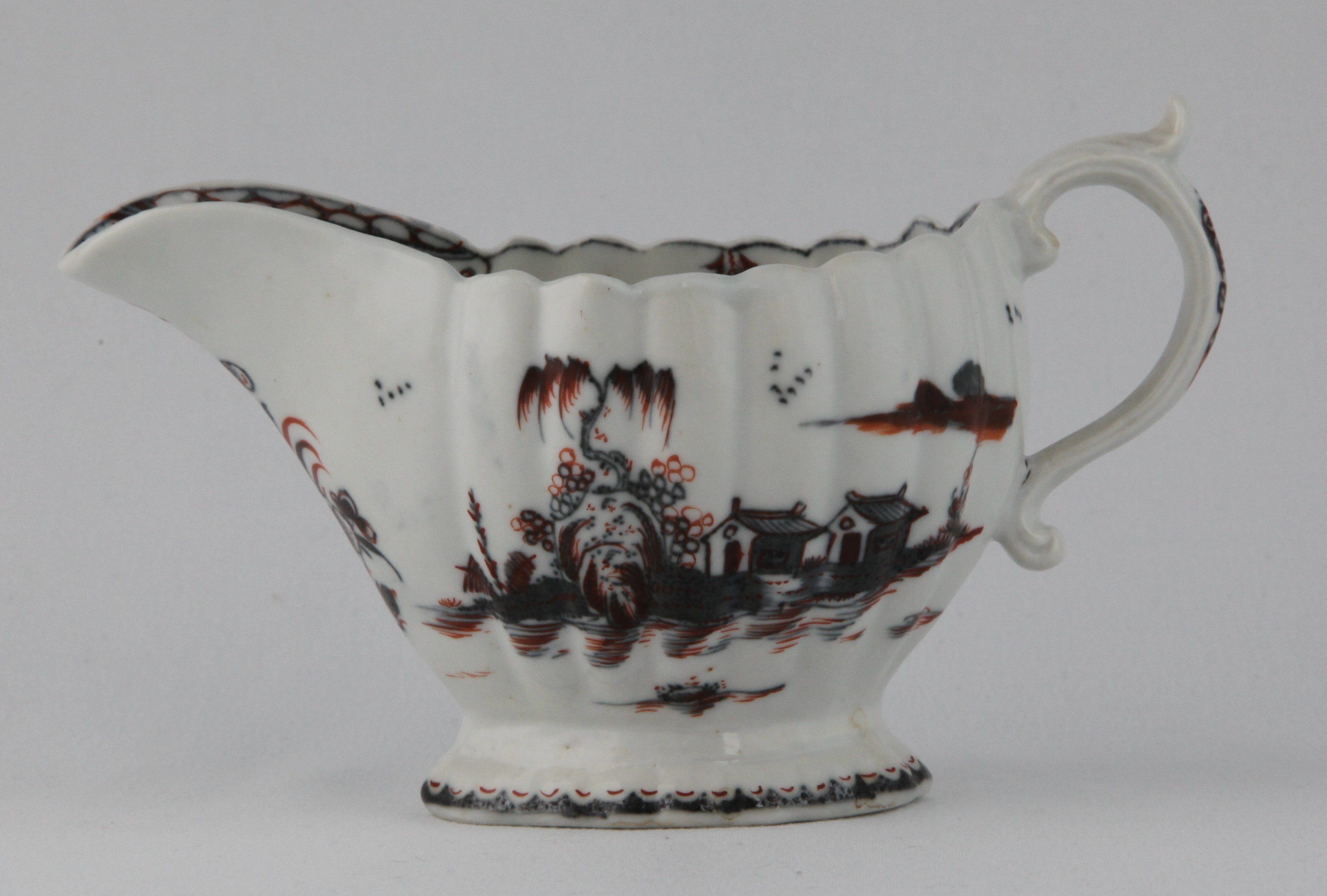 A Plymouth fluted oval sauceboat, the 'S' scroll handle with leaf moulded thumb piece, painted in underglaze blue in Chinese style with buildings in river landscapes, and picked out in iron red enamel, the interior with a flower spray, and pendant flower border, 6 1/8" long, circa 1770