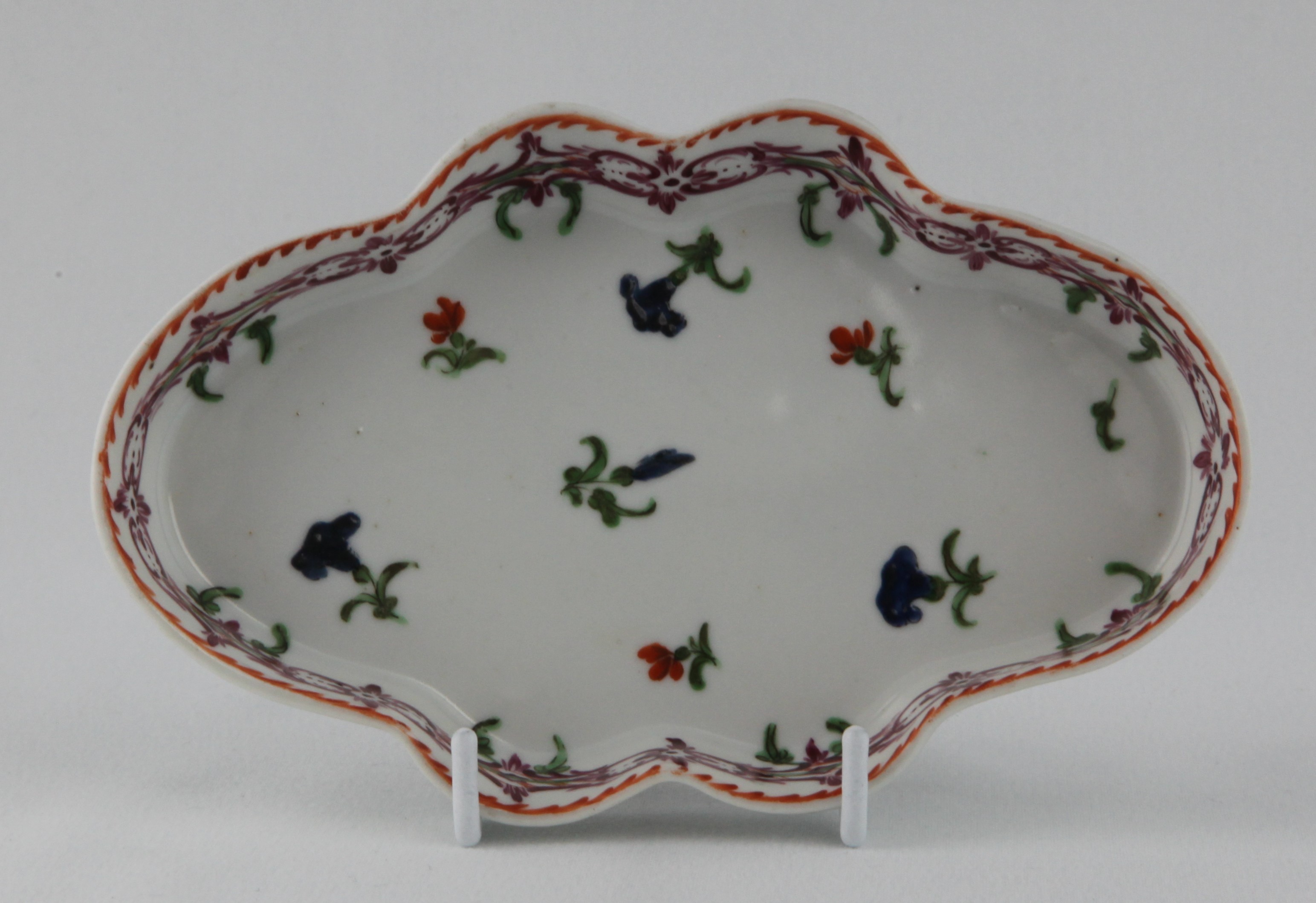 A rare Bristol lobed oval spoon tray, painted in coloured enamels with flower sprigs, within a puce interlinked flower head band, suspending green leaves, and orange angular dentil rim, 6" wide, circa 1775, no mark